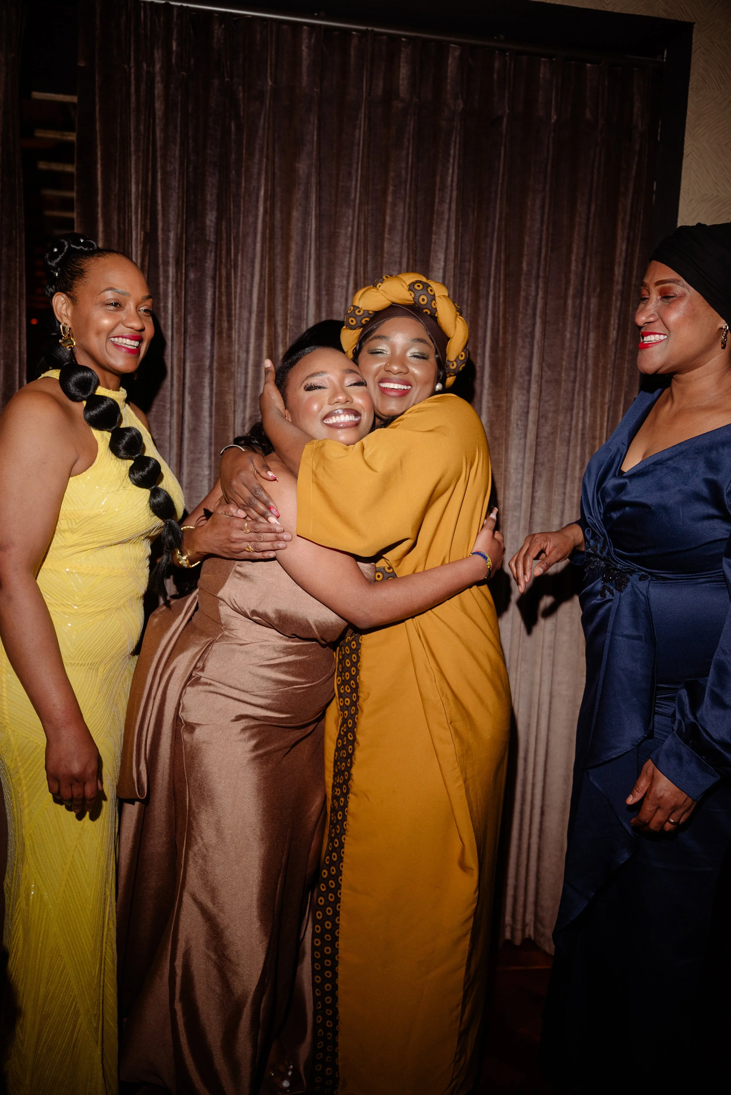 Four women hugging and smiling in a warmly lit room, dressed in colorful evening wear, standing in front of a brown curtain.