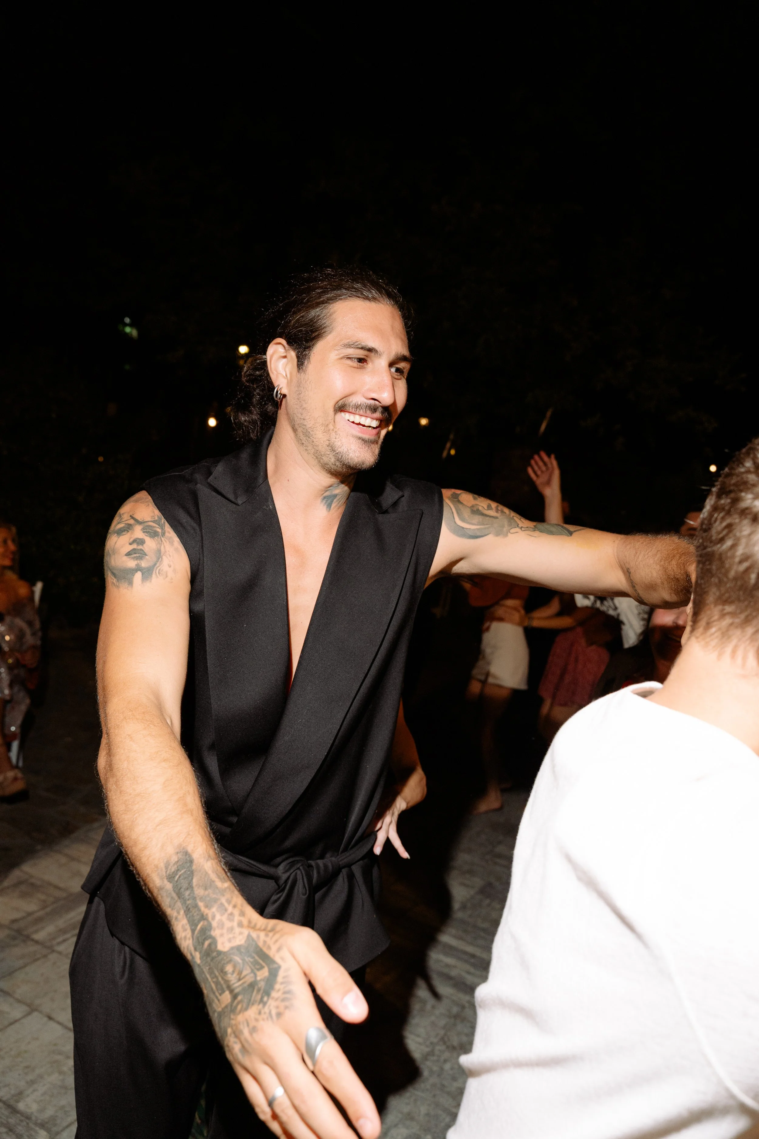 A man with tattoos, wearing a sleeveless black outfit, smiling and dancing at an outdoor nighttime event.