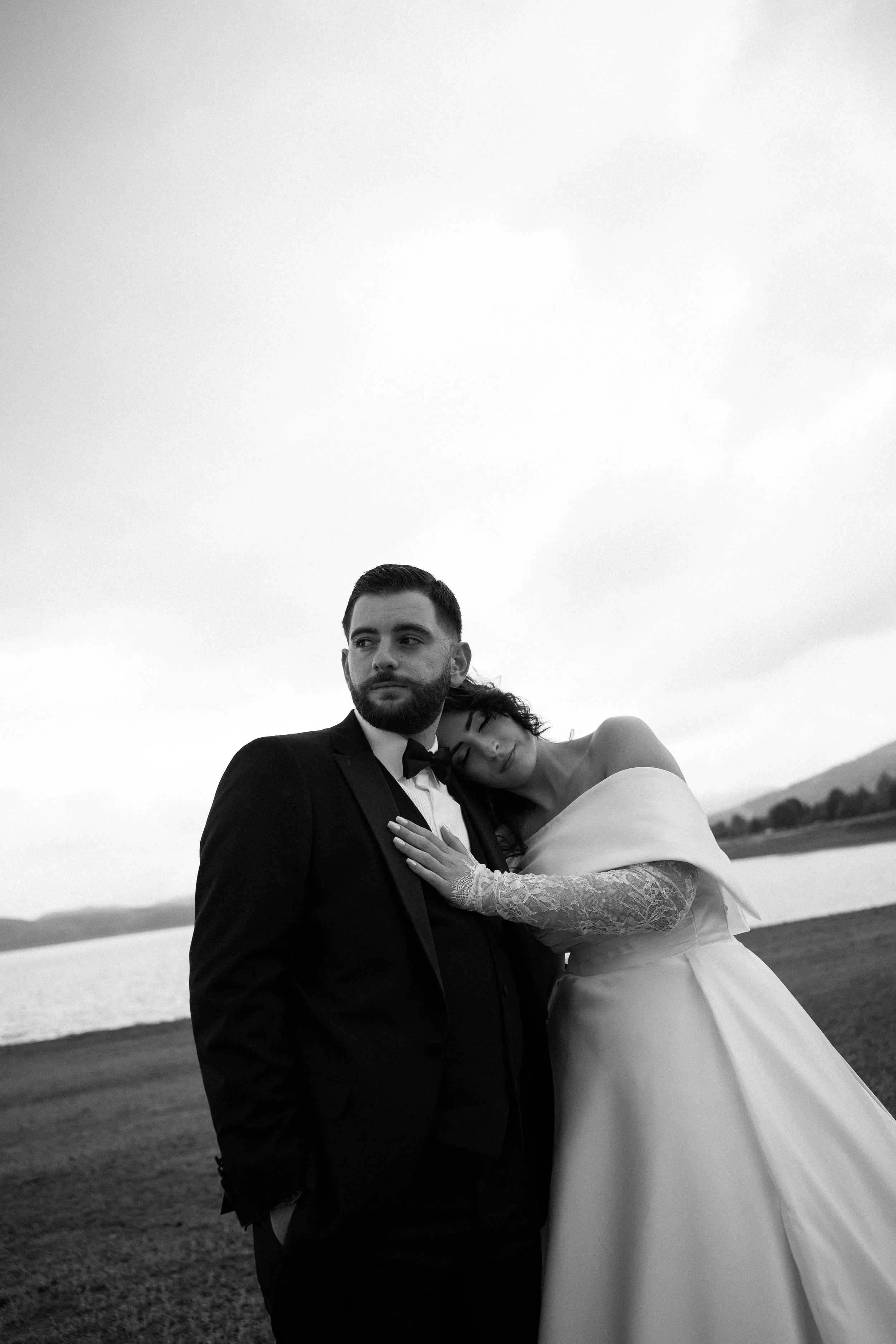 Black and white photo of a bride and groom standing outdoors near a lake, with mountains in the background. The bride, with her eyes closed, rests her head on the groom's shoulder. She is wearing a wedding dress with lace sleeves, and he is wearing a