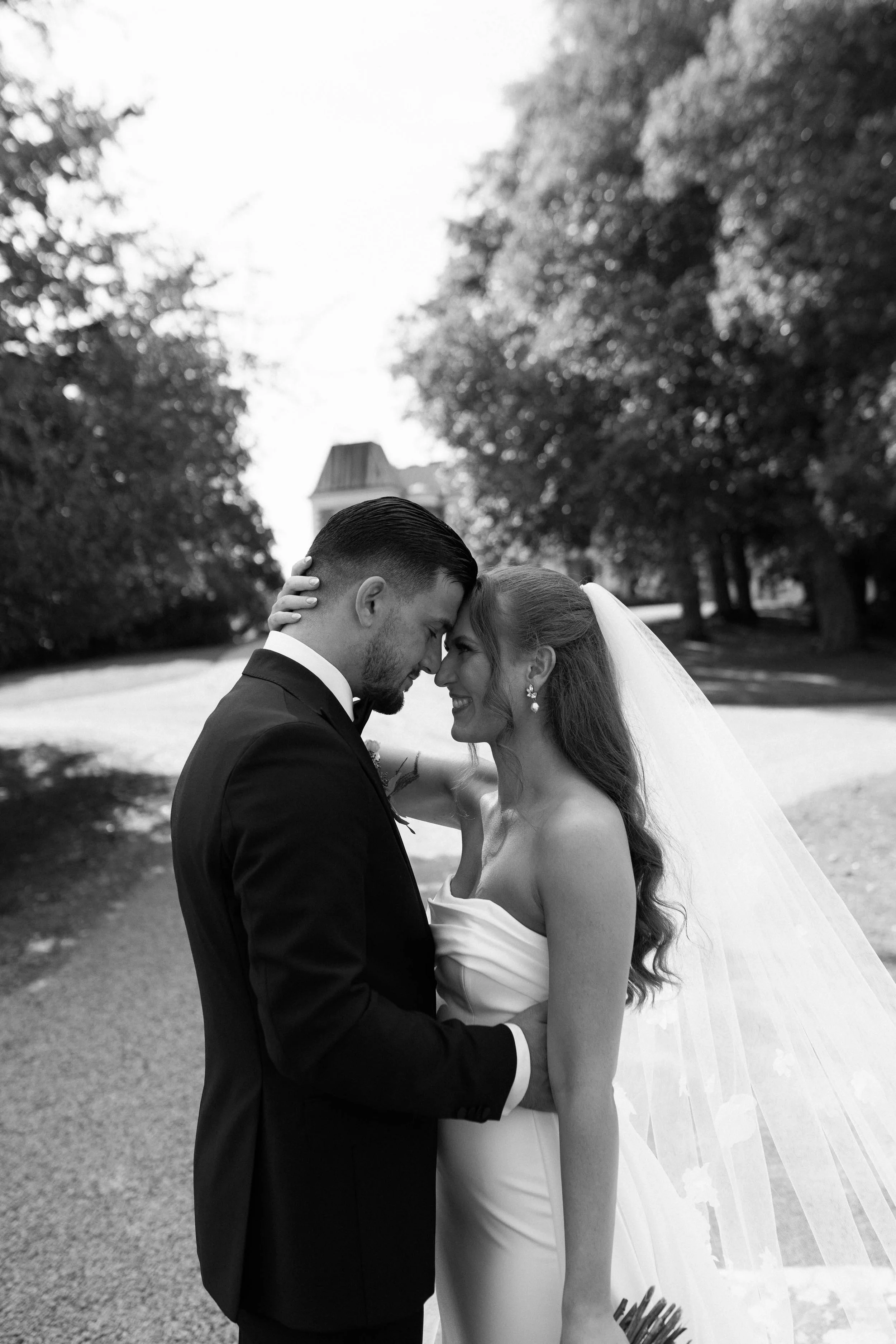 Black and white photograph of a newlywed couple, a man in a tuxedo and a woman in a wedding dress with a veil, standing close with foreheads touching in a park with trees and a building in the background.