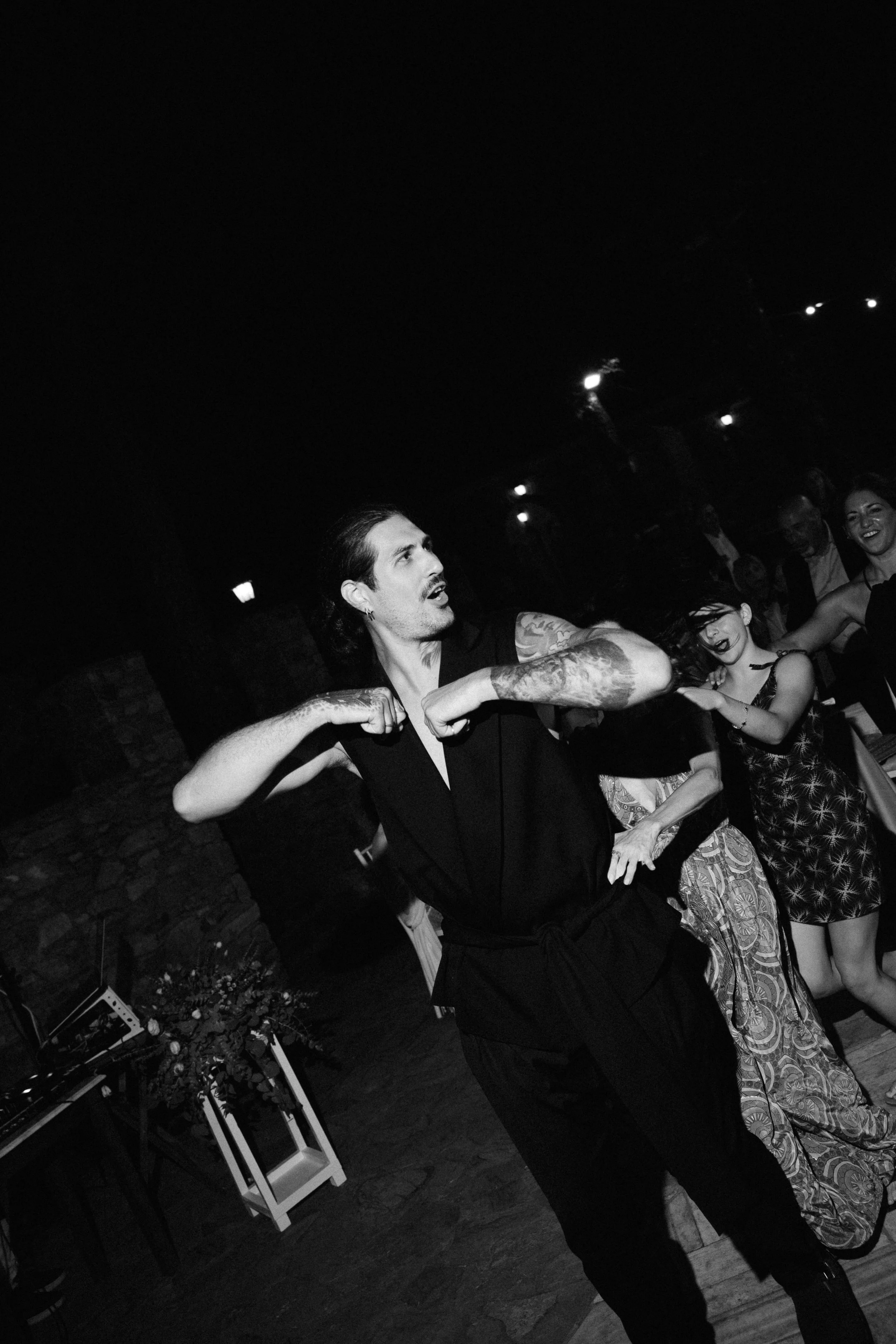 A man with tattoos dancing at a party or celebration with a group of people, black and white photo.