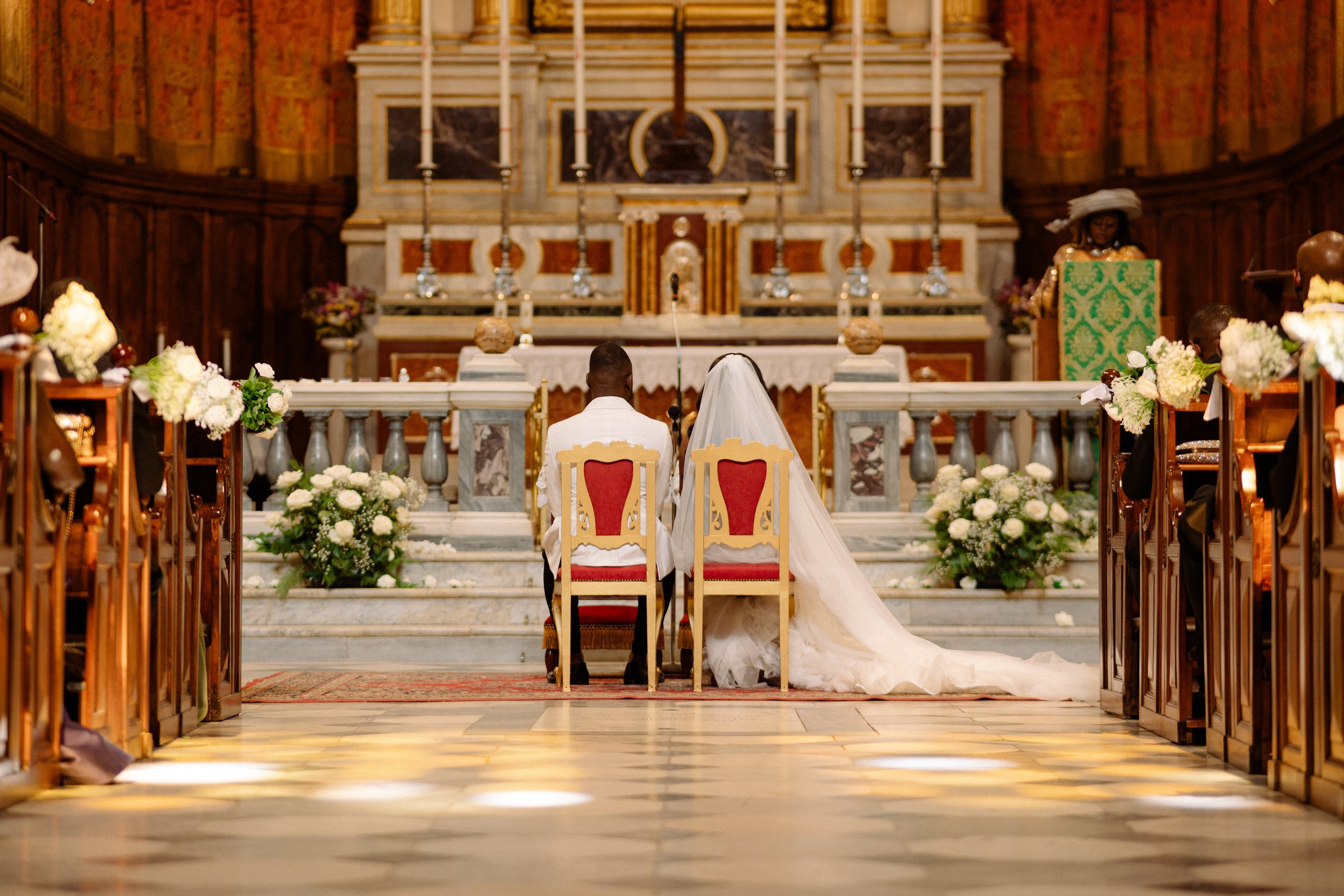 A bride and groom seated before altar during wedding ceremony in a church, surrounded by floral arrangements and candles.