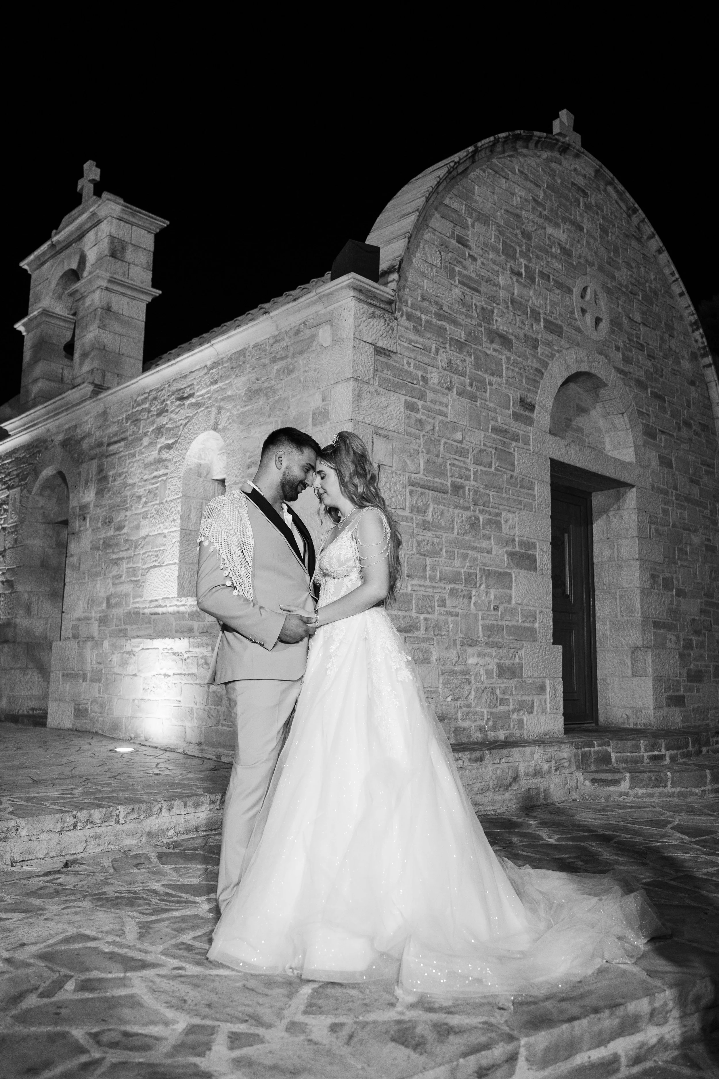 Black and white photo of a bride and groom standing close together in front of a stone building at night, touching foreheads and smiling.