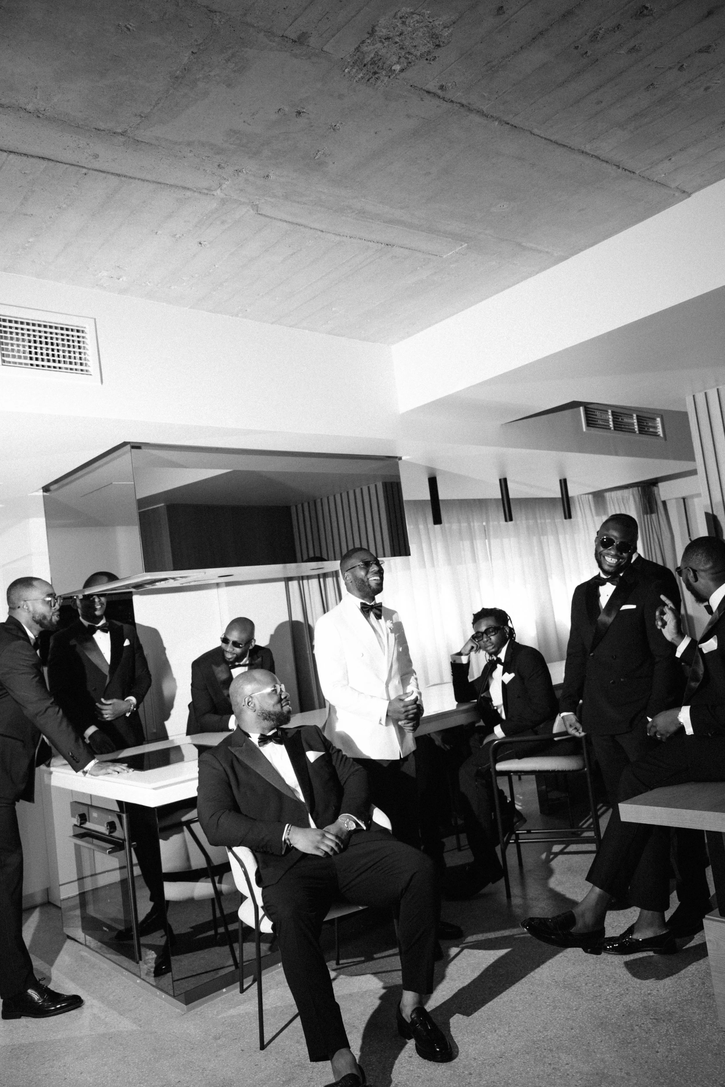 A group of men dressed in tuxedos, some wearing sunglasses, smiles, and laugh in a modern, well-lit indoor space with curtains and contemporary decor.