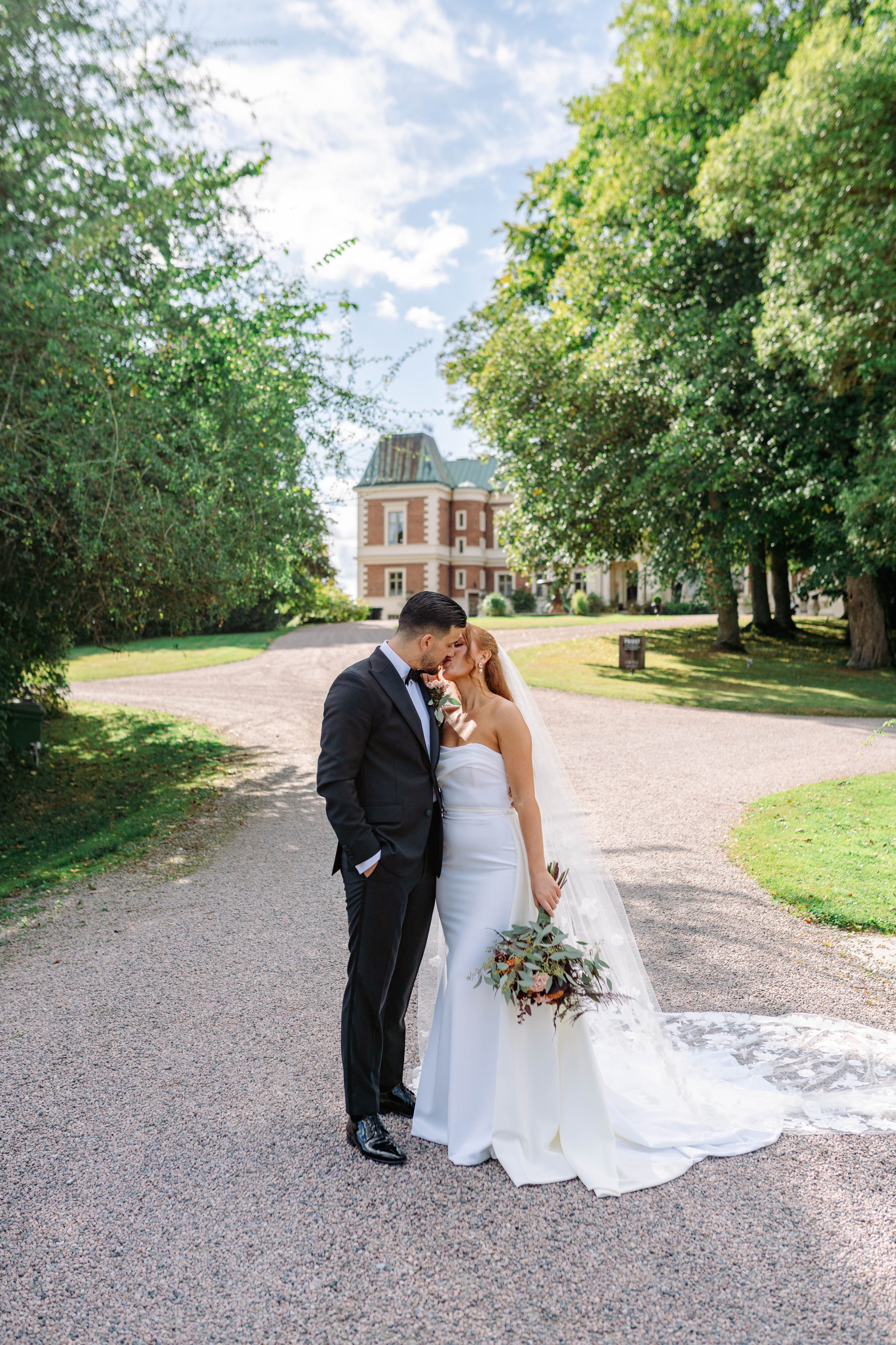 A bride and groom sharing a kiss outdoors on a gravel driveway, surrounded by lush green trees and a large historic house in the background, on a sunny day.