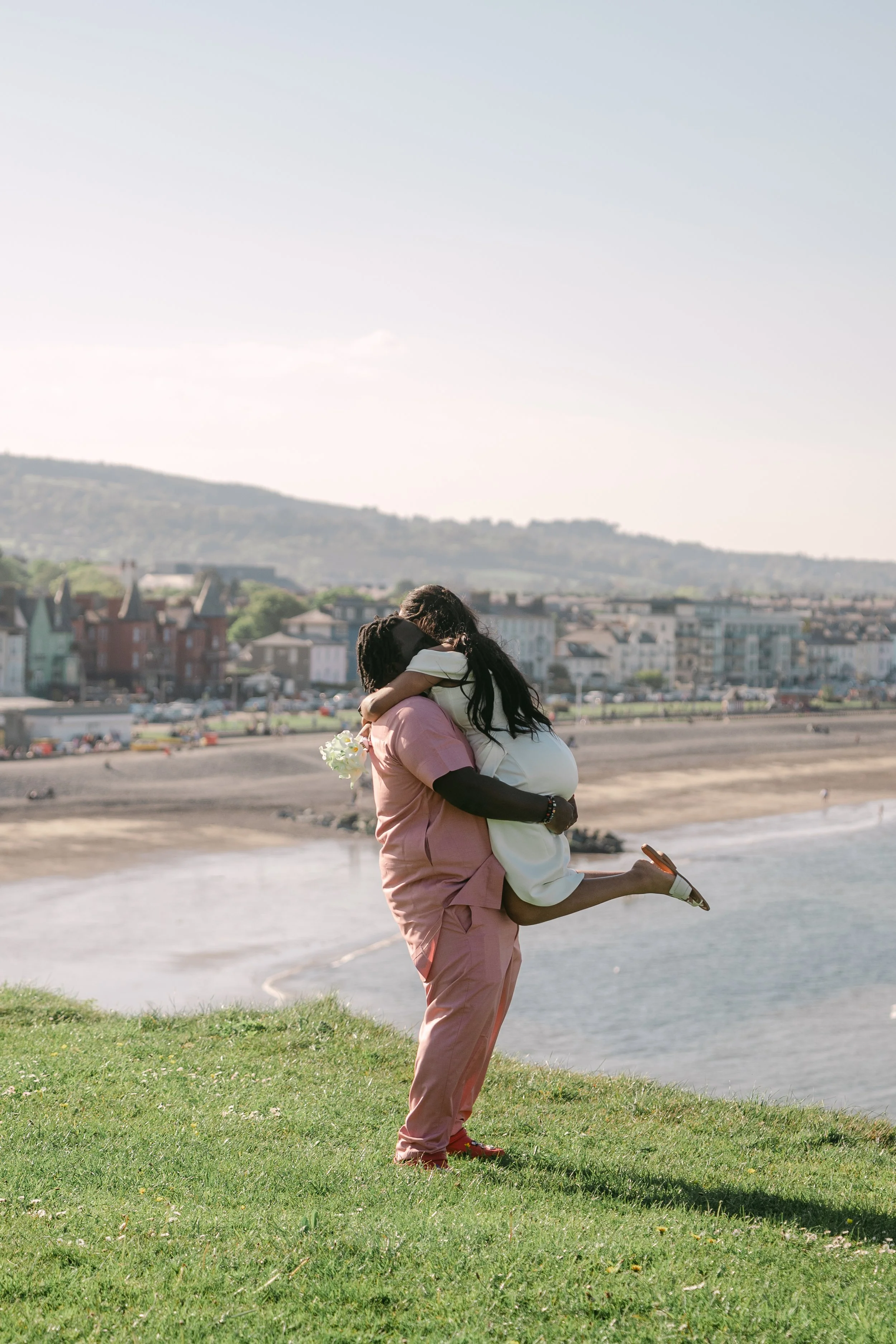 A person in pink scrubs is holding a person in a white dress in a hug on a grassy hill overlooking a beach and a town with colorful houses, under a clear sky.