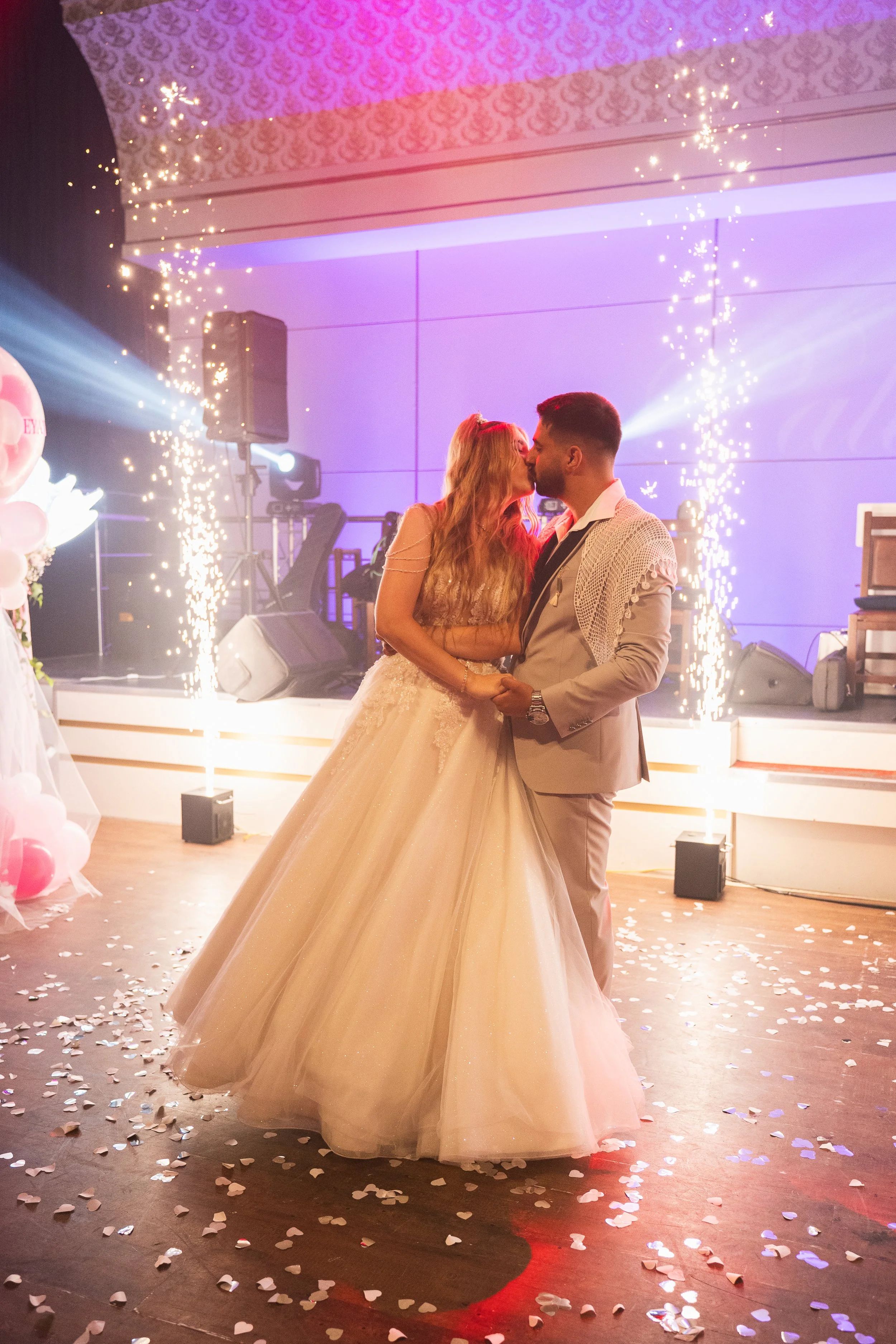 A bride and groom on the dance floor sharing a kiss at their wedding reception, surrounded by fireworks, confetti, and colorful lighting.