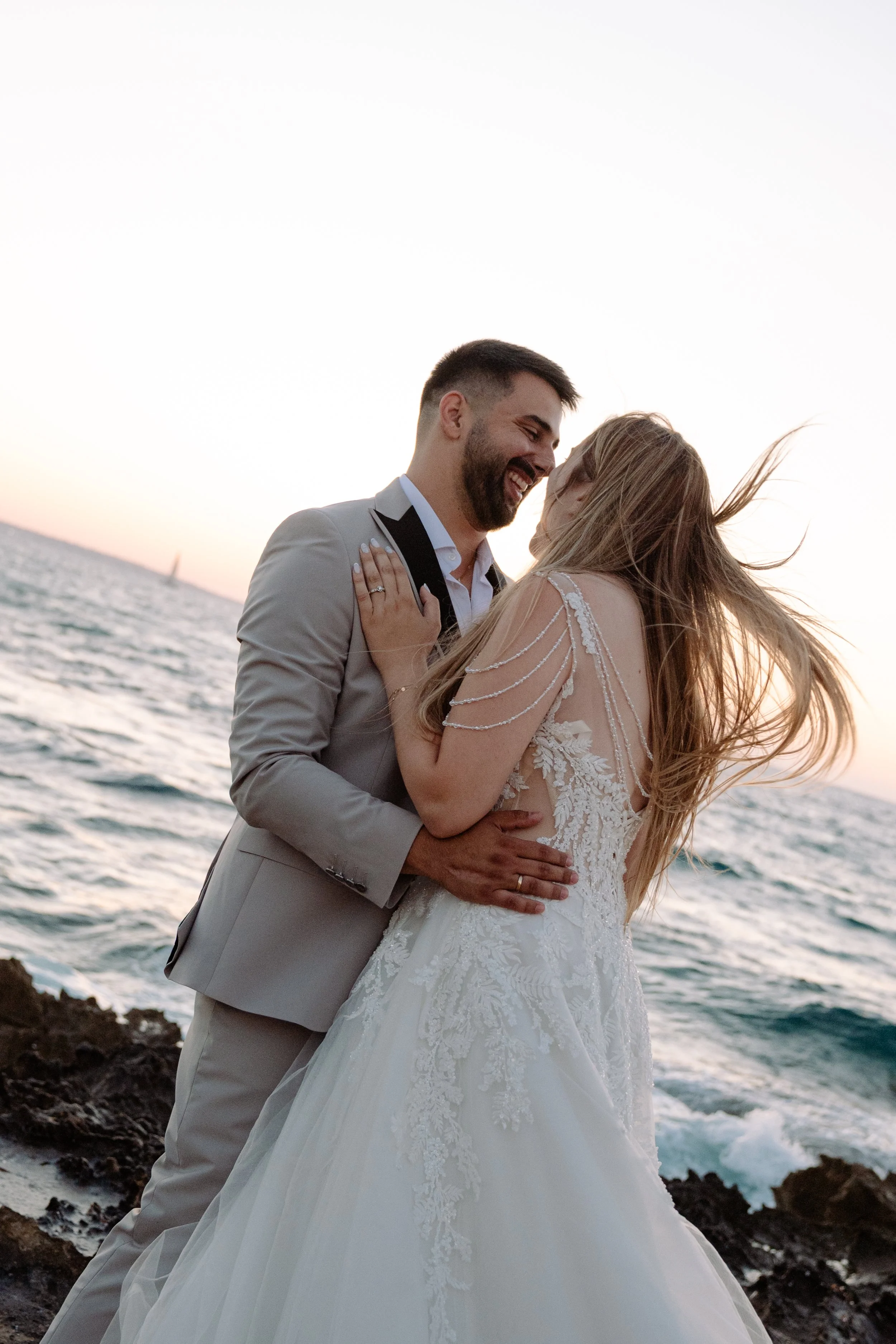 A couple in wedding attire sharing a romantic moment by the water at sunset, with the man wearing a gray suit and the woman in a white lace wedding dress.