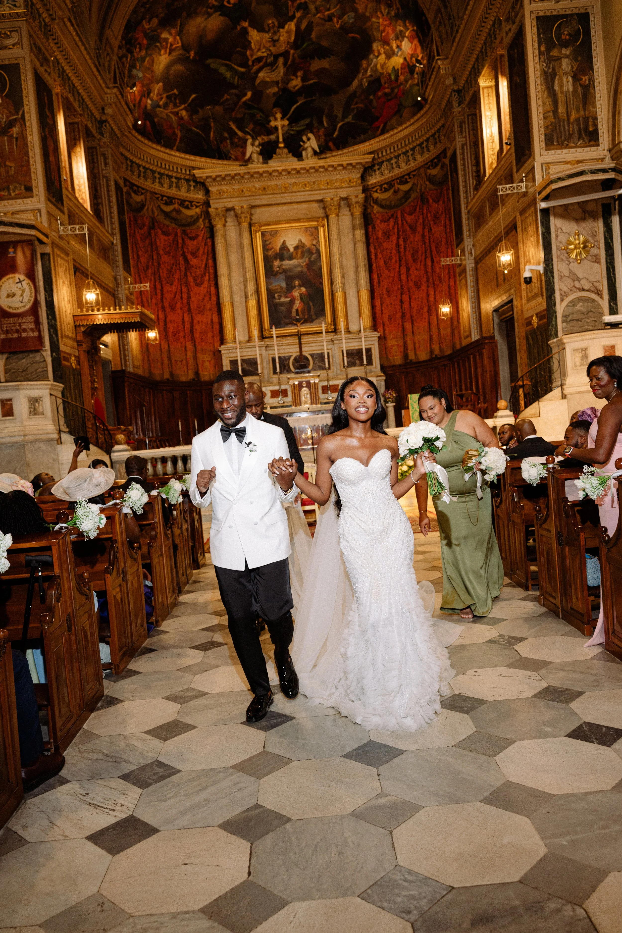 A newlywed couple walking down the aisle of a church, holding hands and smiling. The groom is wearing a white tuxedo jacket with black pants and bow tie, and the bride is in a white lace wedding gown holding a bouquet of white roses. Guests are seate