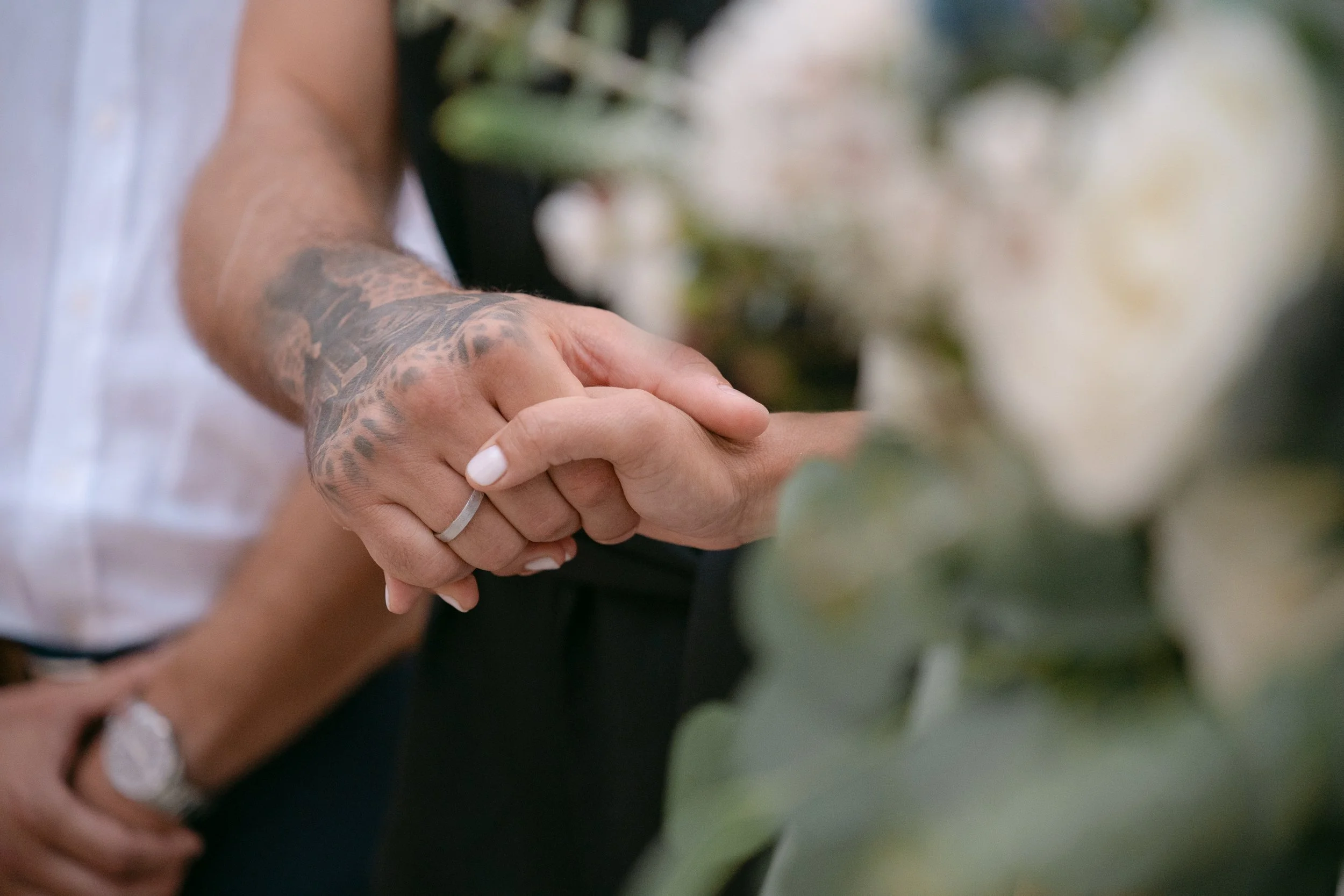 Two people holding hands, one with a tattooed arm and a ring on the finger, while the other has a clean, unmarked hand, near a blurred background of white flowers or roses.