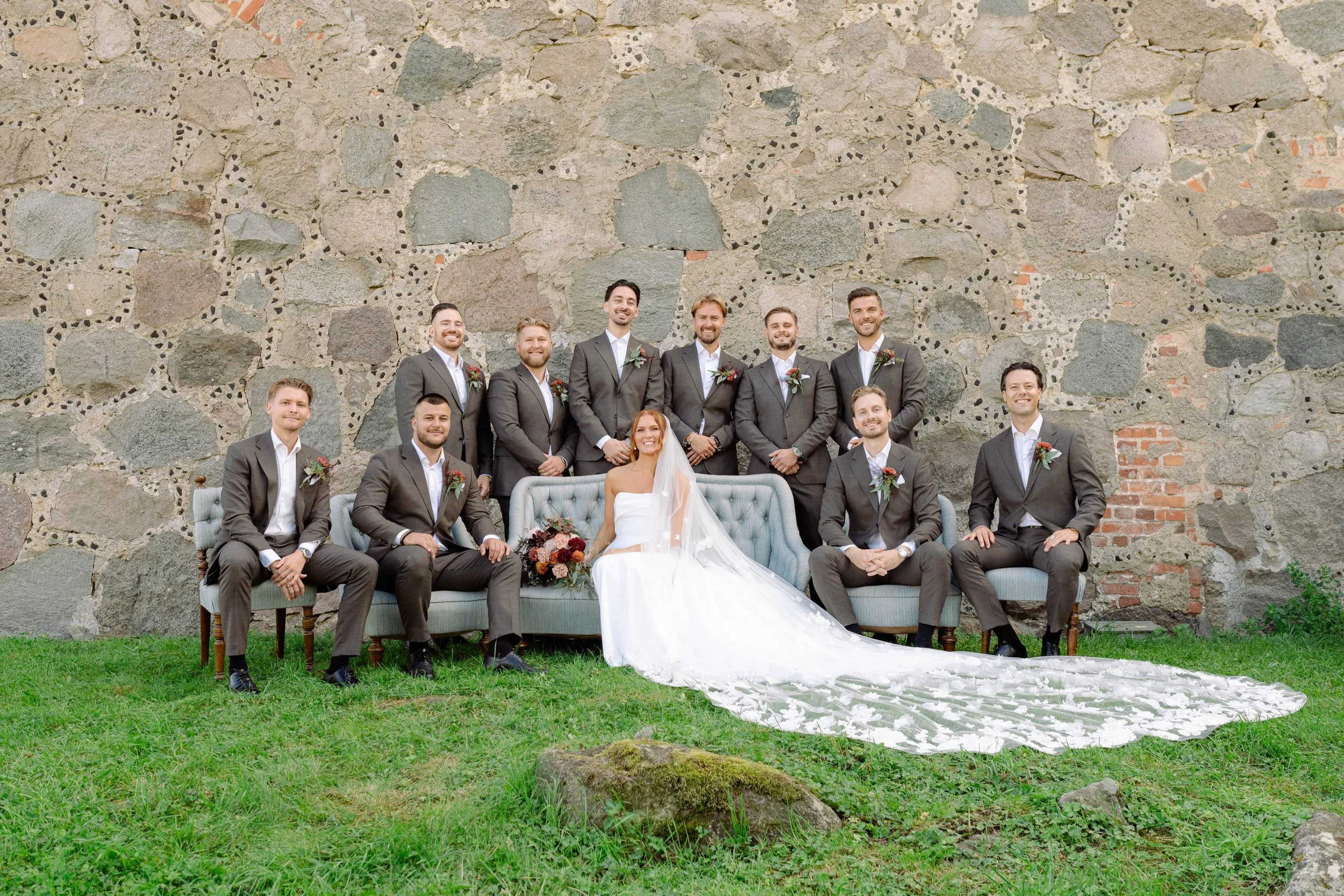 A bride in a white wedding dress with a long, lace train sits on a vintage blue sofa outdoors, surrounded by her wedding party. The groom and groomsmen in gray suits with boutonnières stand and sit around her against a stone wall, smiling.
