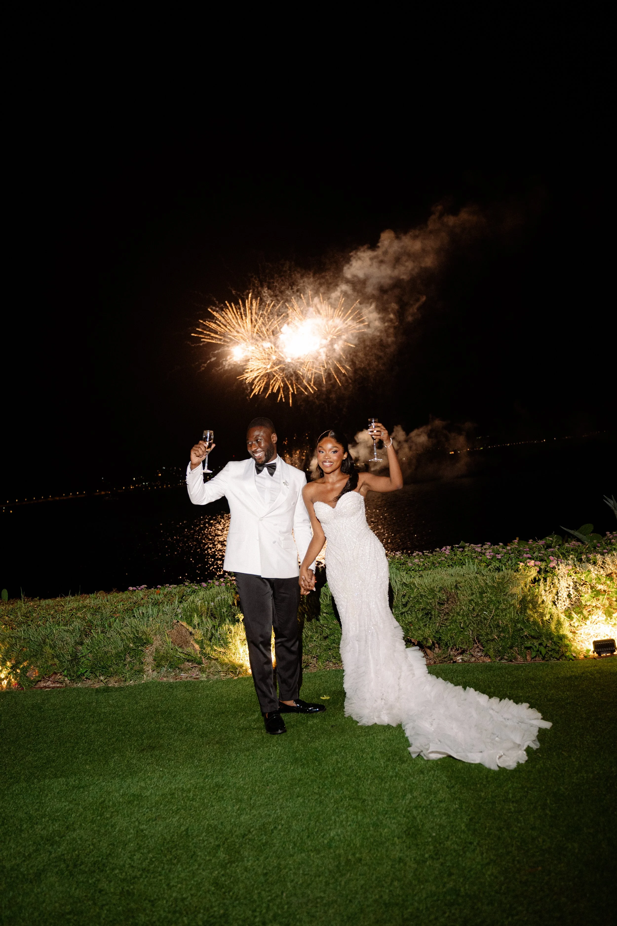 A newlywed couple celebrating outdoors at night by a body of water with fireworks in the sky, holding glasses of champagne and smiling.