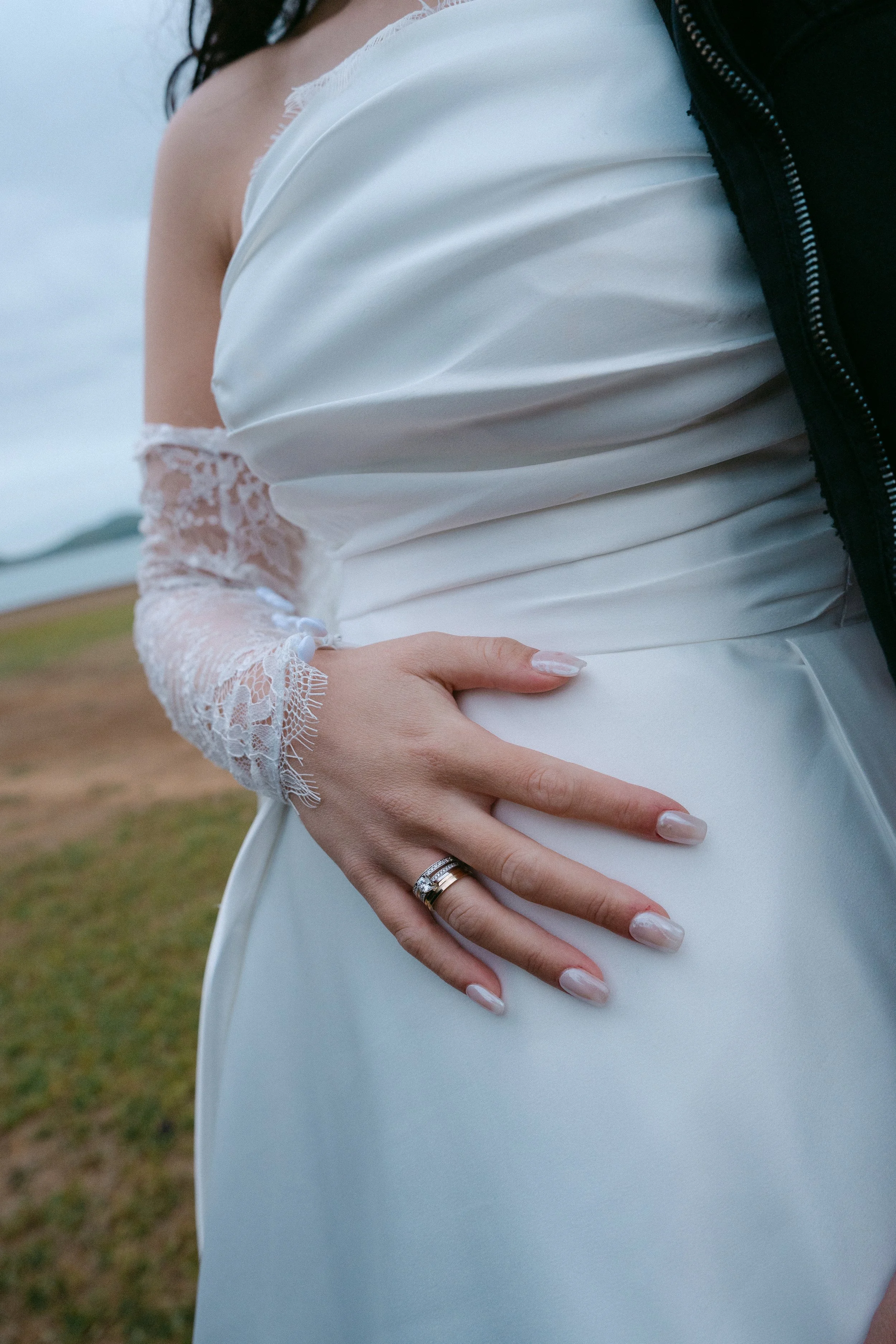 Close-up of a woman wearing a white dress with lace sleeves, holding her pregnant belly outdoors, with a cloudy sky in the background.