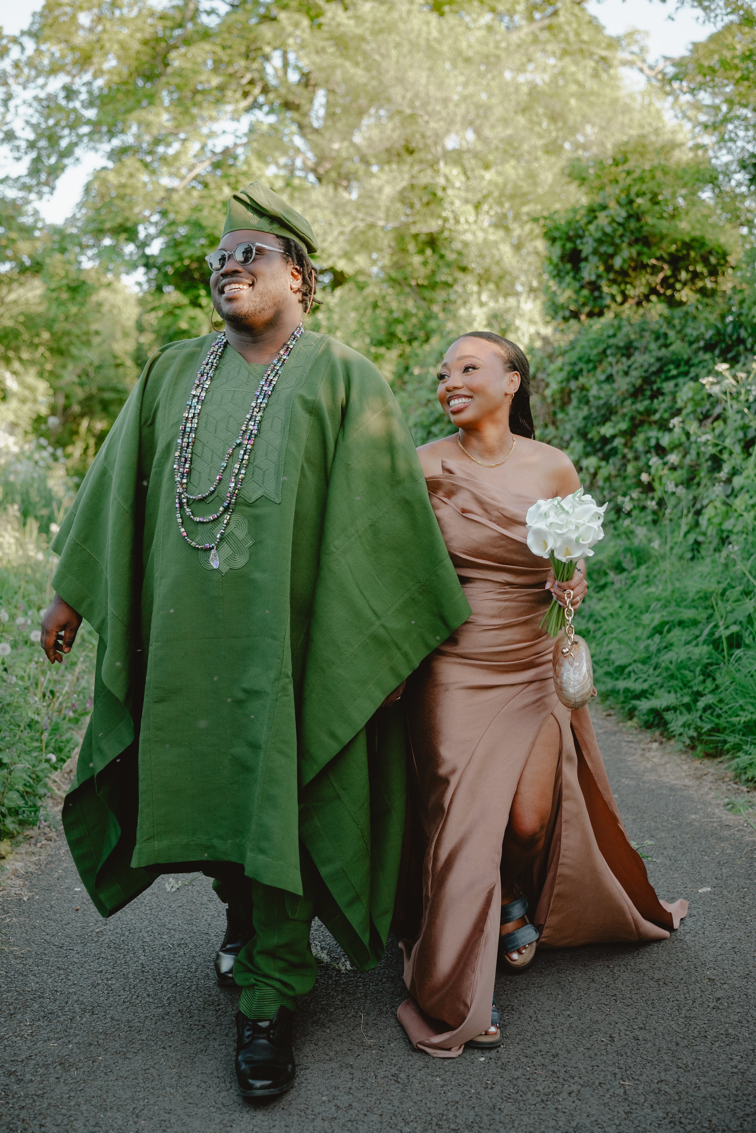 A couple dressed in formal attire walking outdoors on a paved path, surrounded by lush green trees and plants. The man is wearing a traditional green outfit with a matching hat, sunglasses, and beaded necklaces. The woman is holding a bouquet of whit