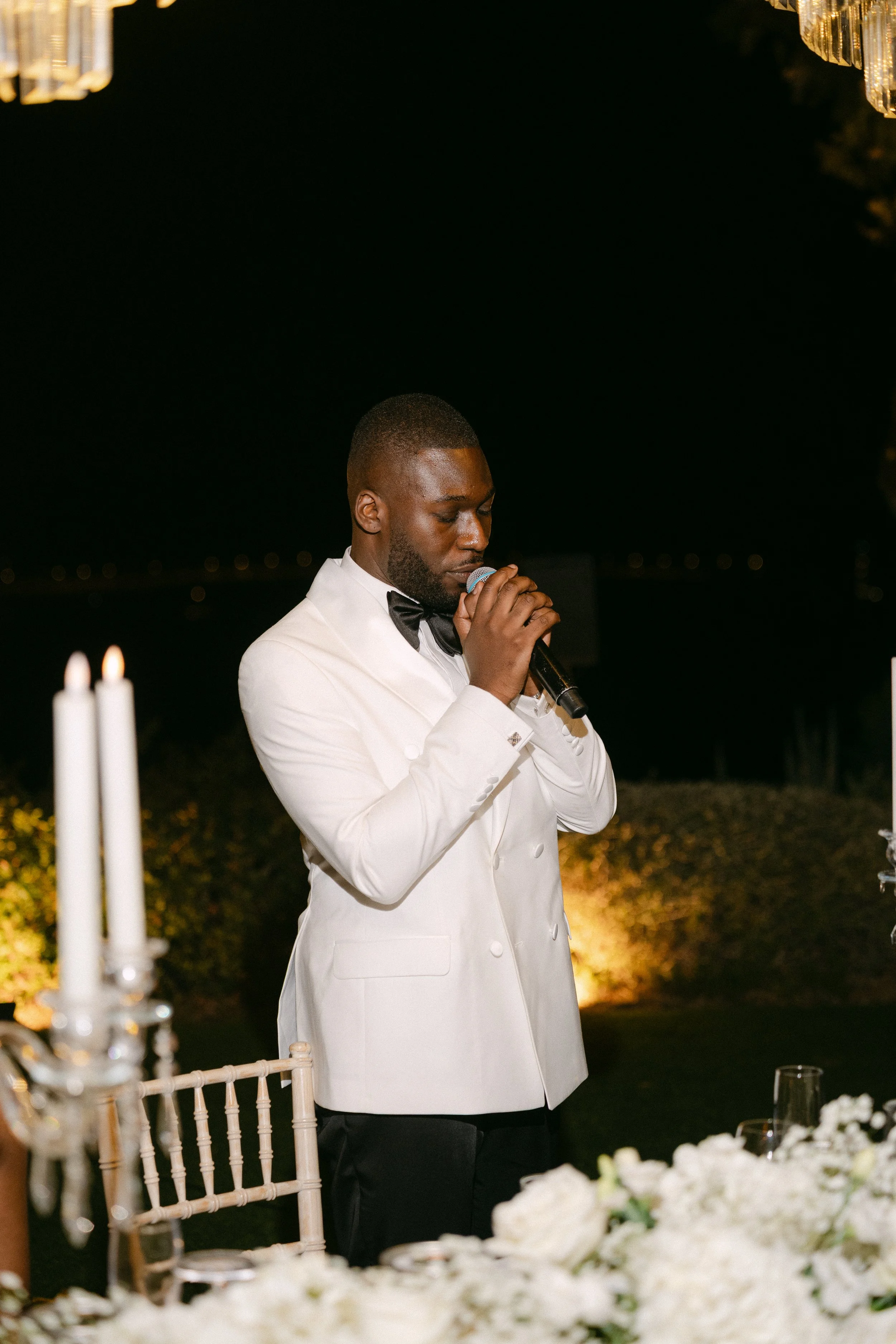 A man in a white tuxedo jacket and black bow tie is holding a microphone and appears to be giving a speech at an outdoor nighttime event, with a table of white flowers and candles in the foreground.