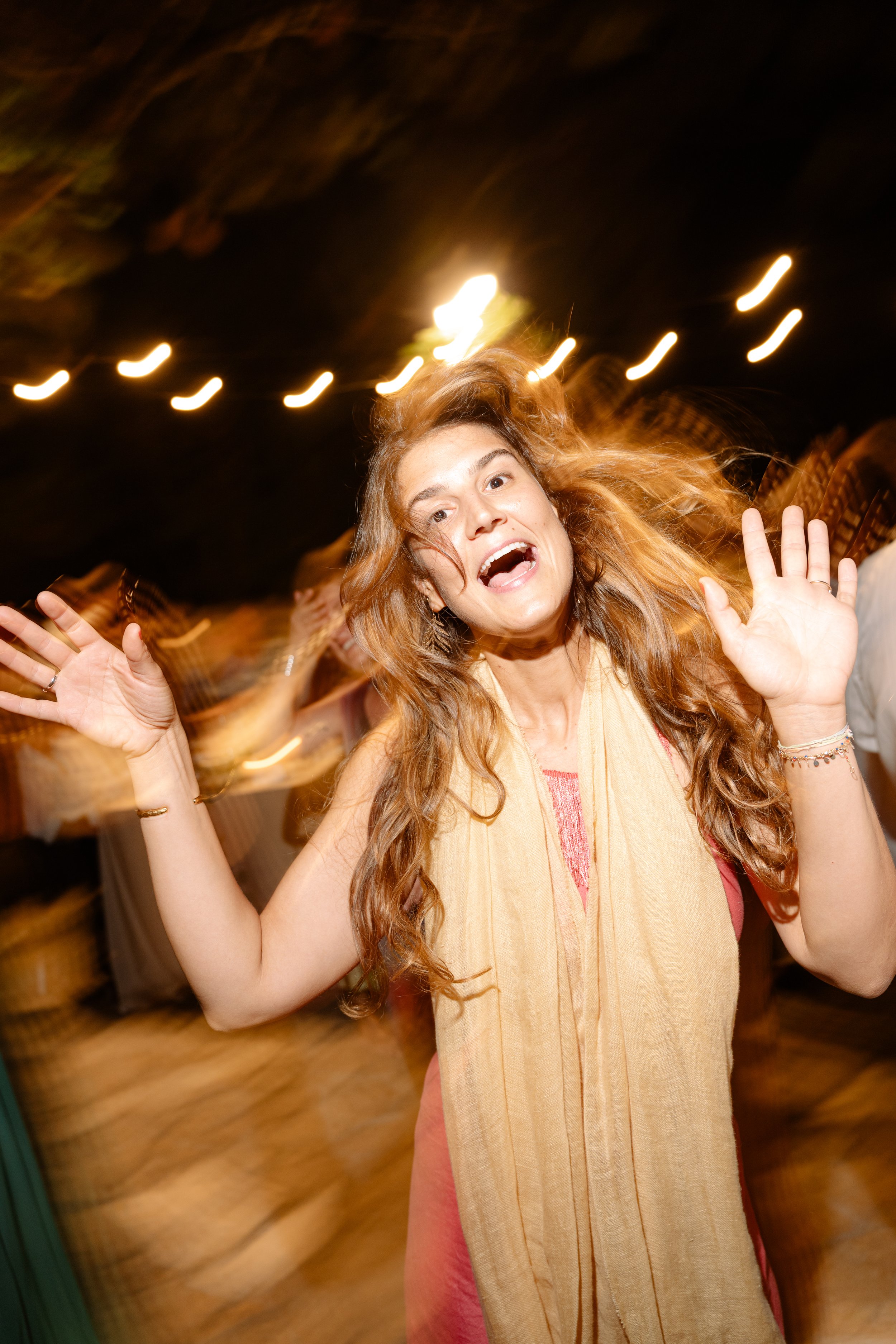 A woman with red hair smiling and waving her hands at night with blurry lights in the background.