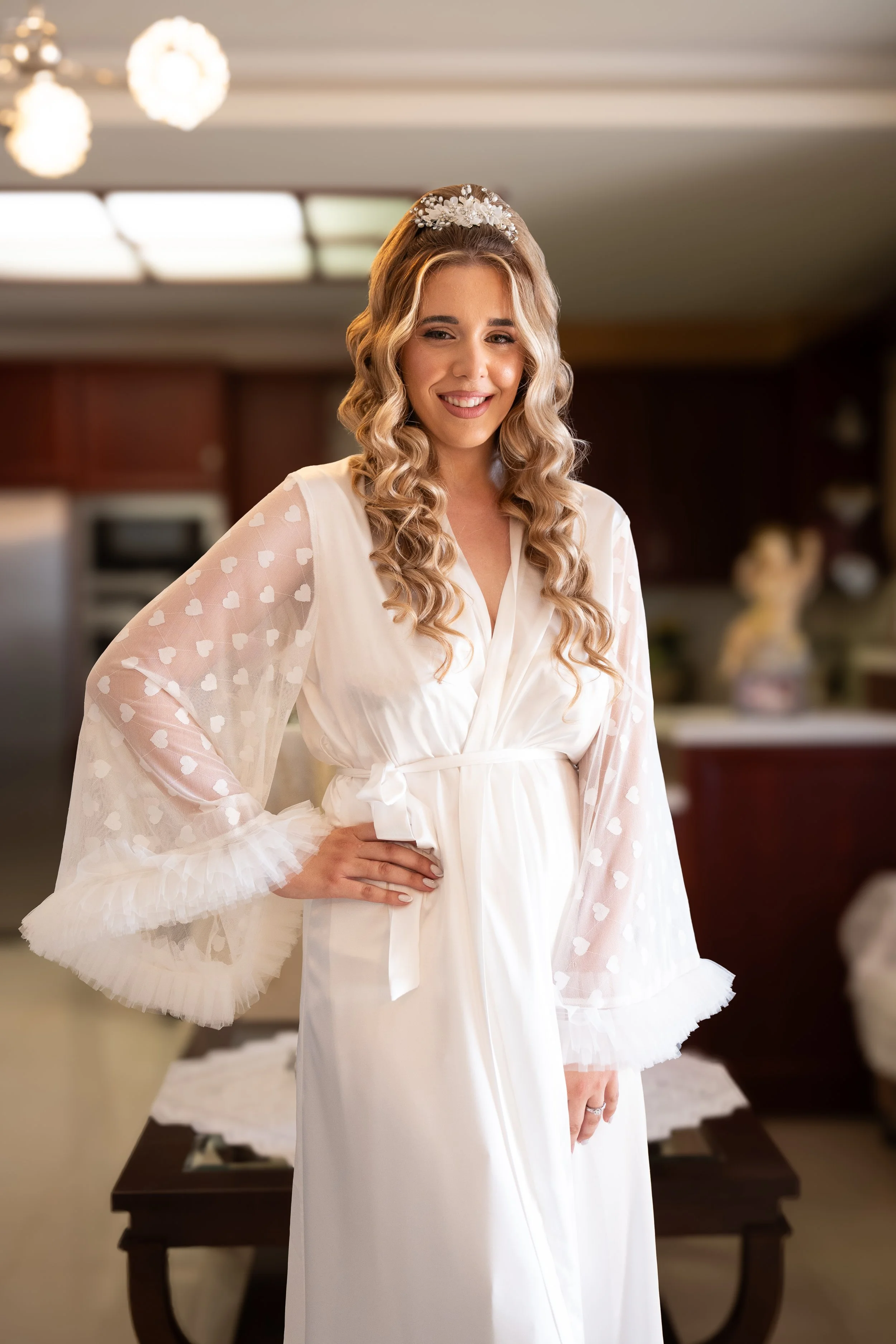 A woman in a white satin robe with sheer, heart-patterned, ruffled sleeves, smiling and standing in a kitchen.