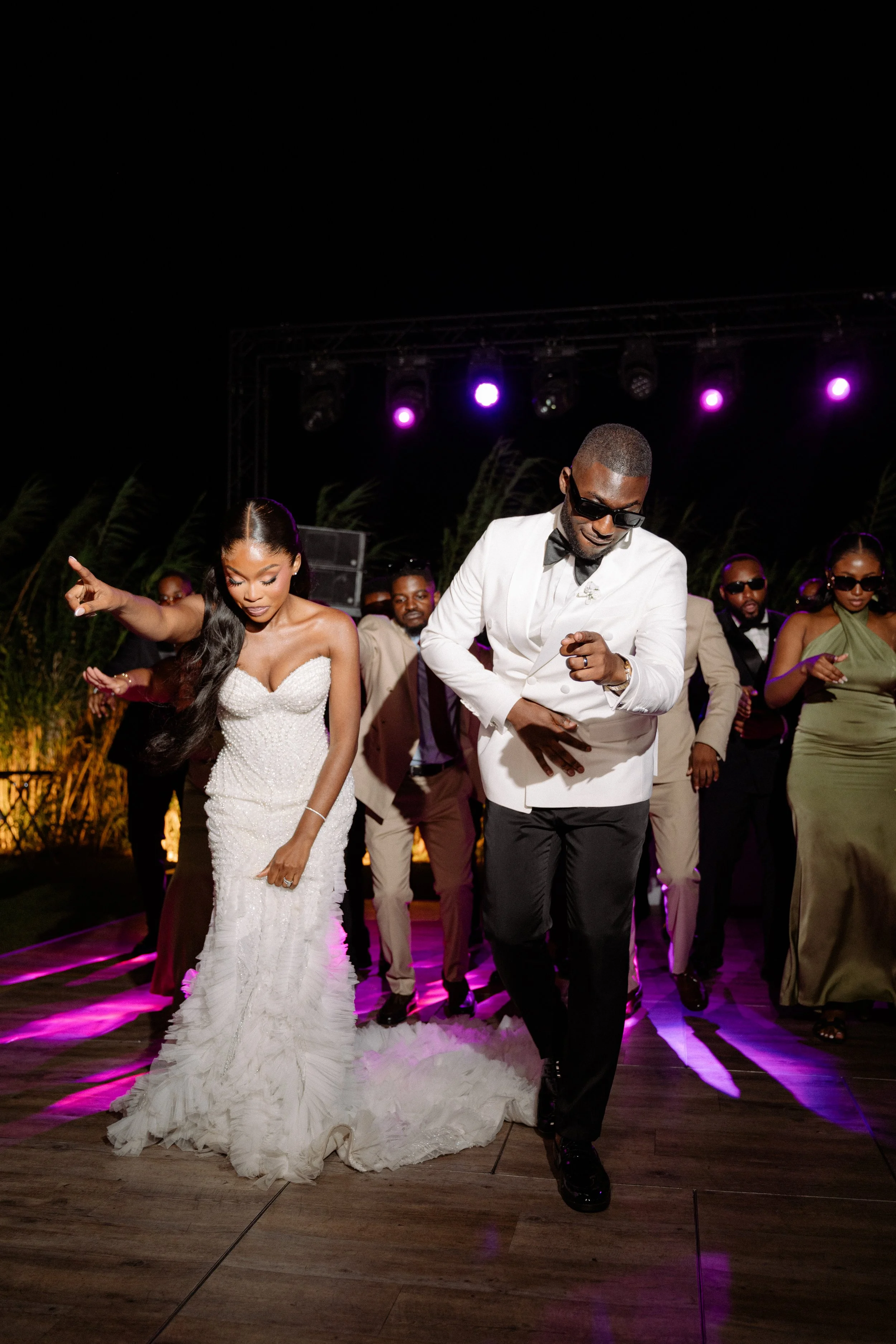 People dancing at a wedding reception during nighttime under purple lights, with the bride in a white gown and the groom in a white tuxedo leading the dance.
