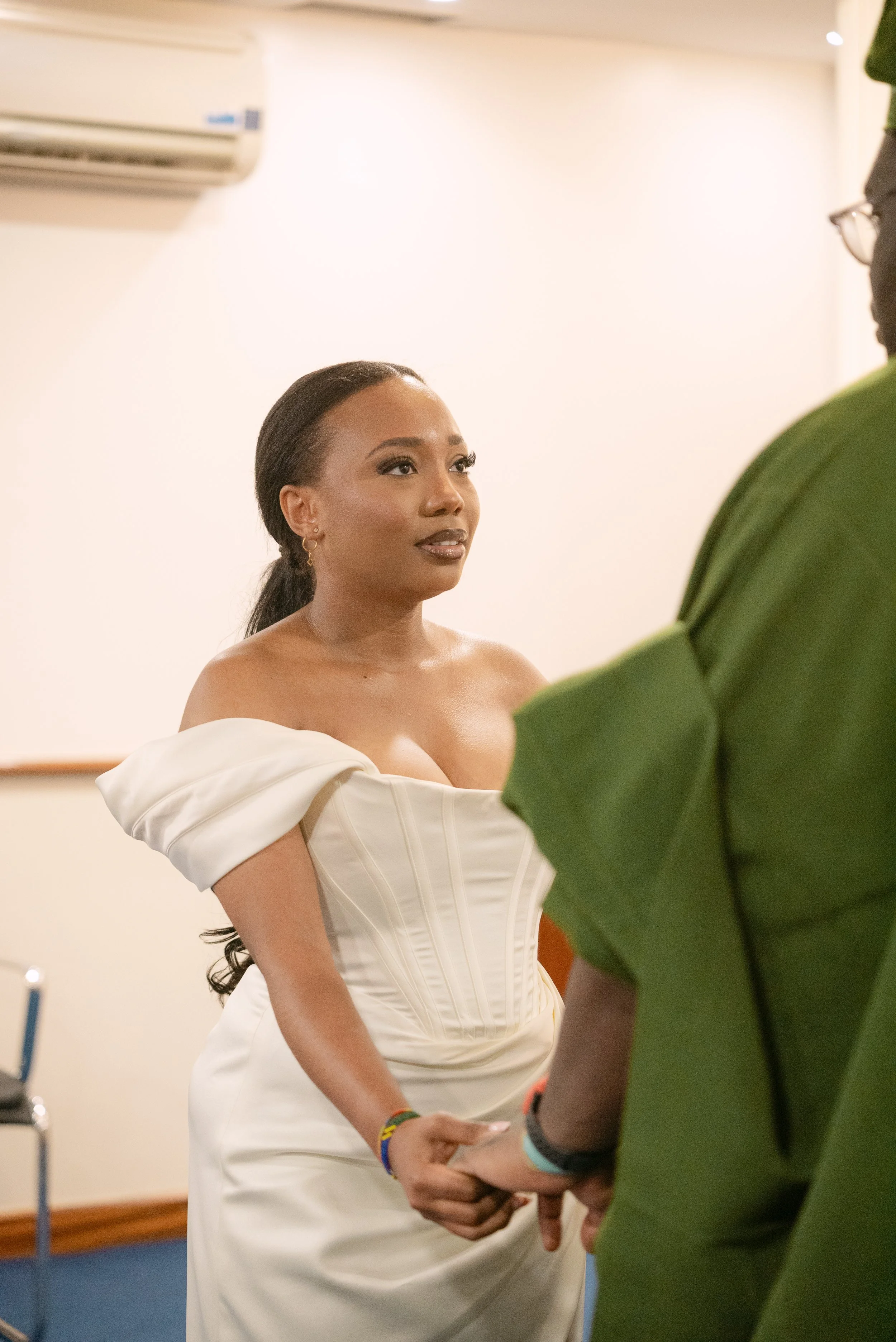 A woman in an off-shoulder white dress holding hands with a person in a green shirt during a wedding or formal ceremony.