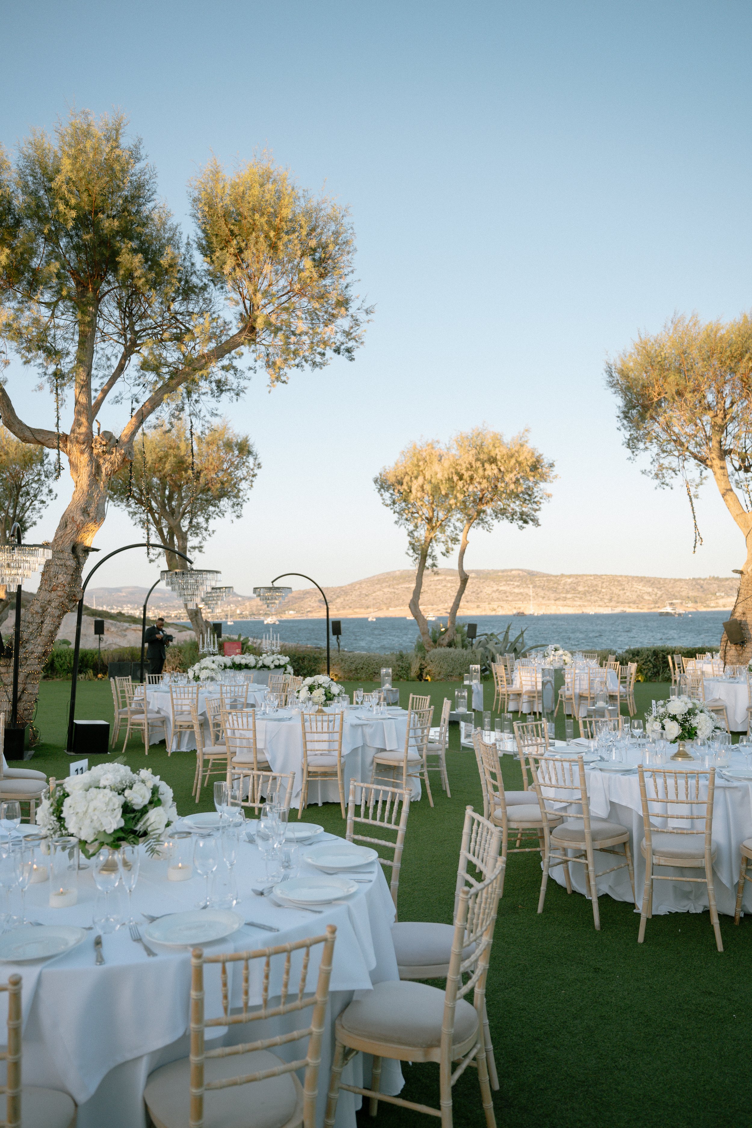 Outdoor wedding reception setup with round tables covered with white tablecloths, floral centerpieces, and white chairs, near the water with trees and hills in the background.