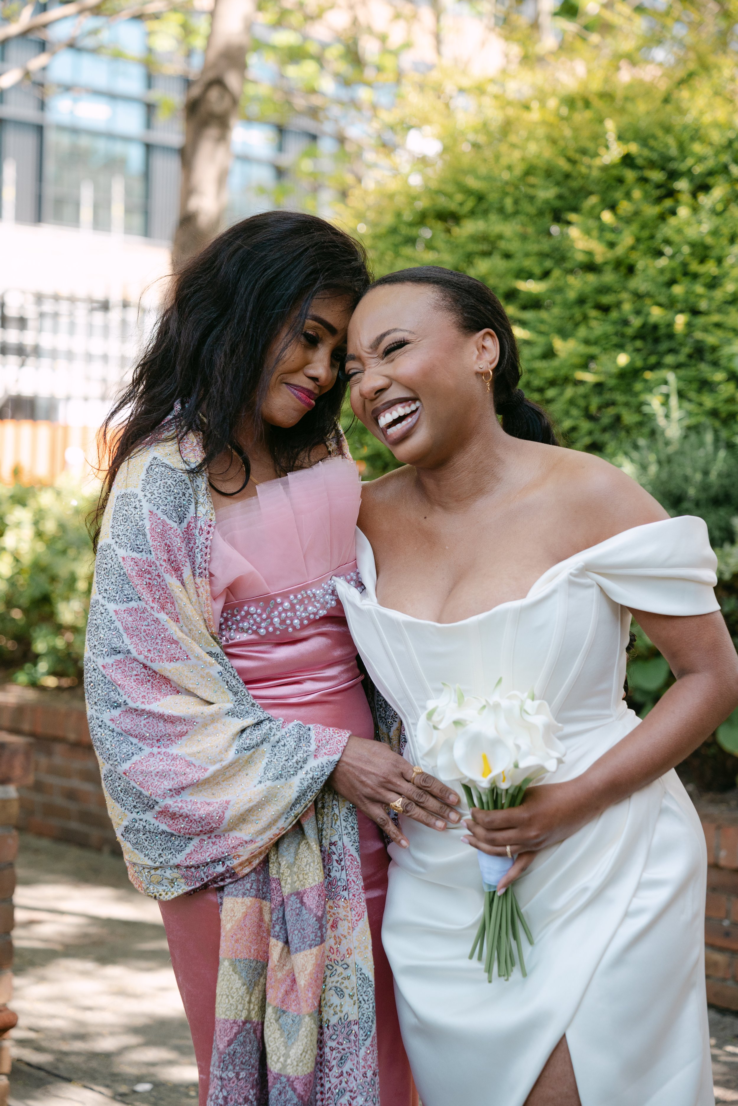 Two women sharing a joyful moment outdoors; one in a white bridal dress holding a bouquet of white calla lilies, the other in colorful clothing with a pink dress, both smiling and touching foreheads.