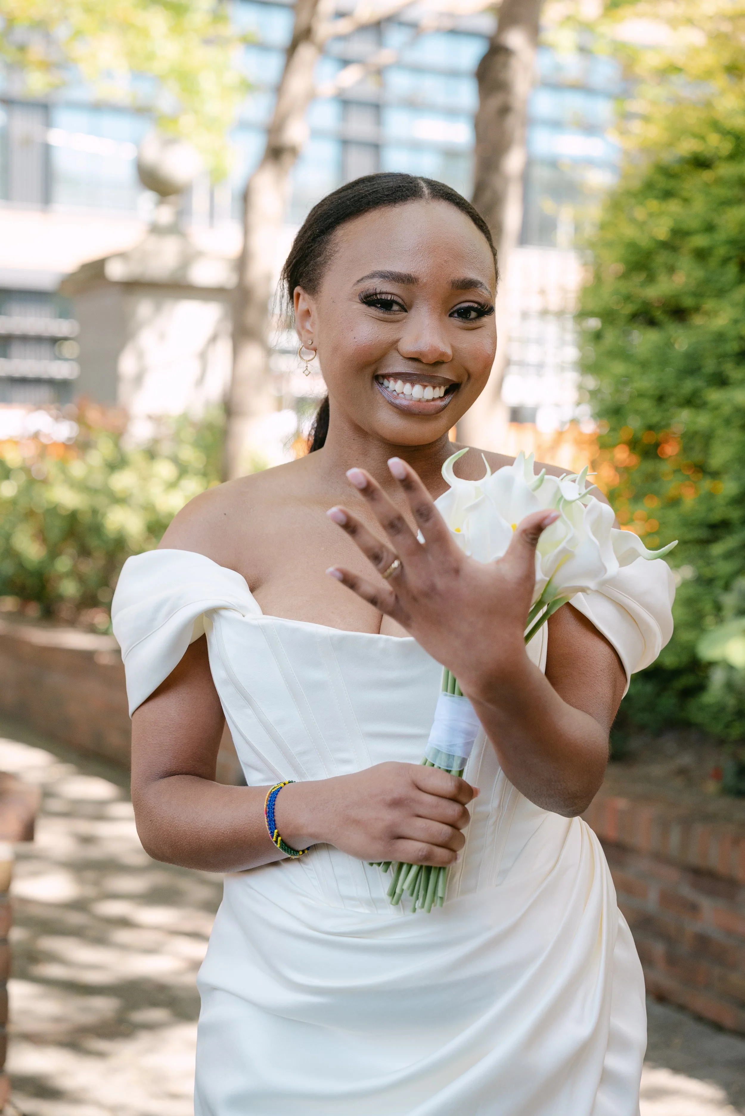 A smiling woman in a white wedding dress holding a bouquet of white calla lilies outdoors.