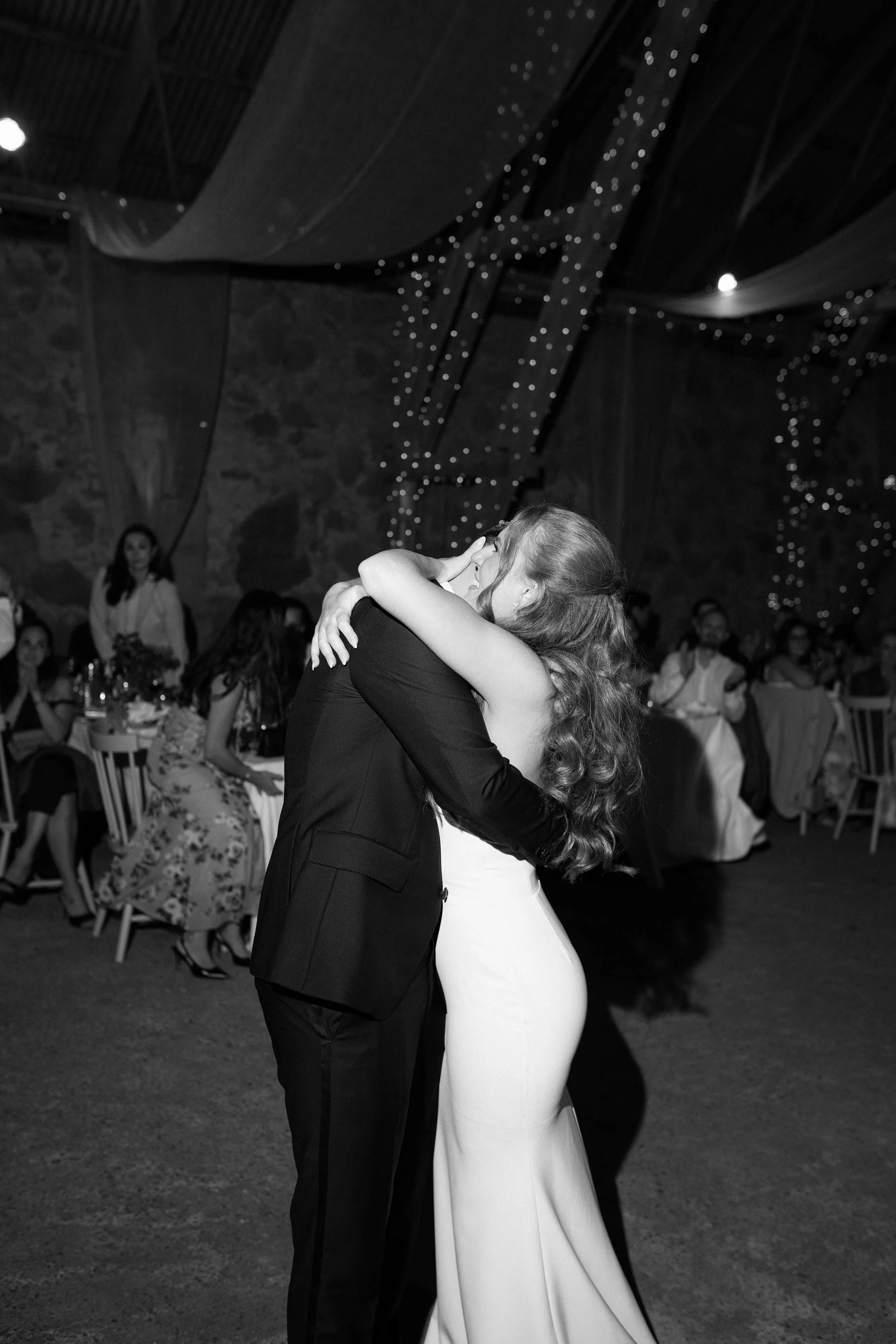 A couple dancing and embracing at a wedding reception, with guests seated at tables watching in the background.