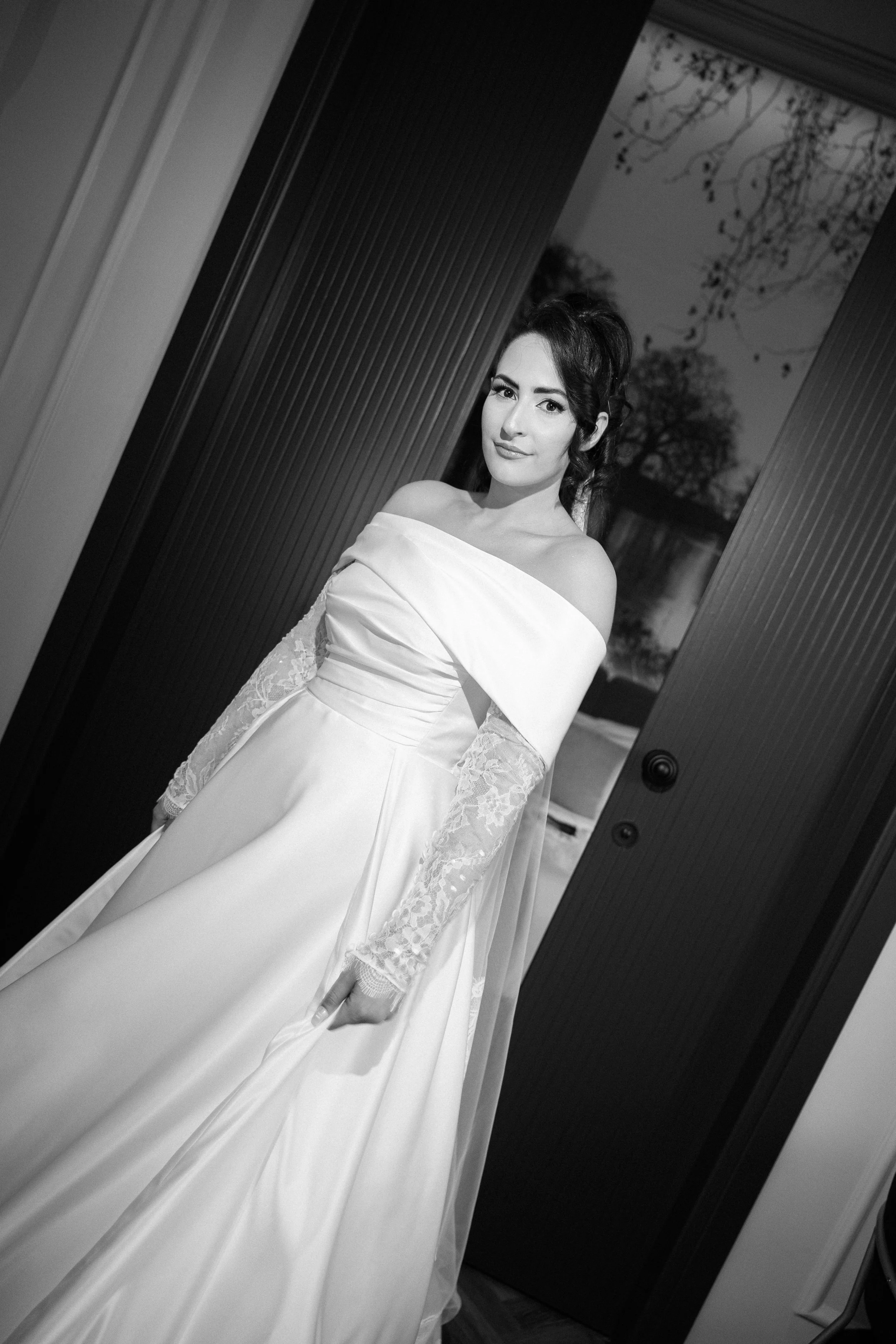 A woman in a white off-the-shoulder wedding dress with lace sleeves, standing inside and looking at the camera, with a door and outdoor scene in the background.