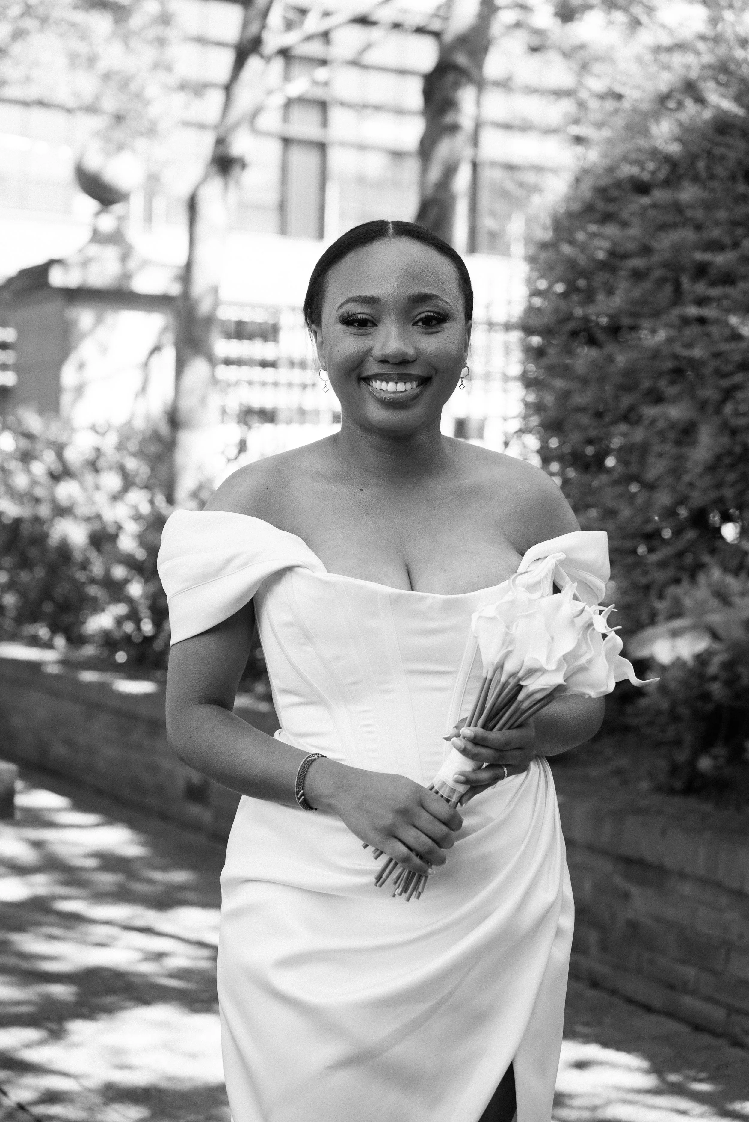 A woman in a white off-shoulder dress holding a bouquet of calla lilies, smiling outdoors in a garden with trees and a brick wall.