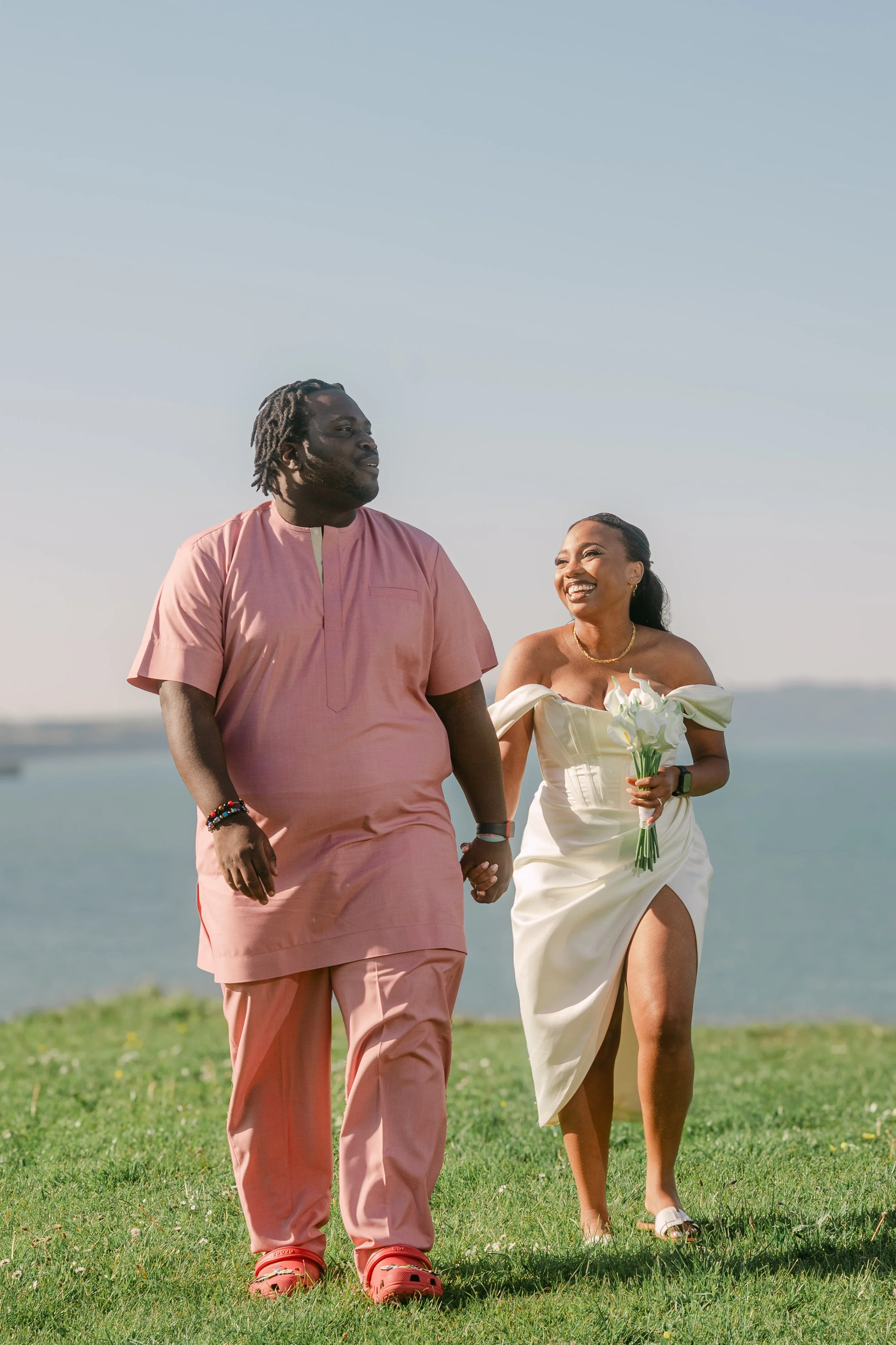 A couple walking outdoors near a body of water, holding hands and smiling. The woman is wearing a white dress with a thigh-high slit and holding a bouquet of white flowers. The man is wearing a light pink traditional outfit and red sandals.