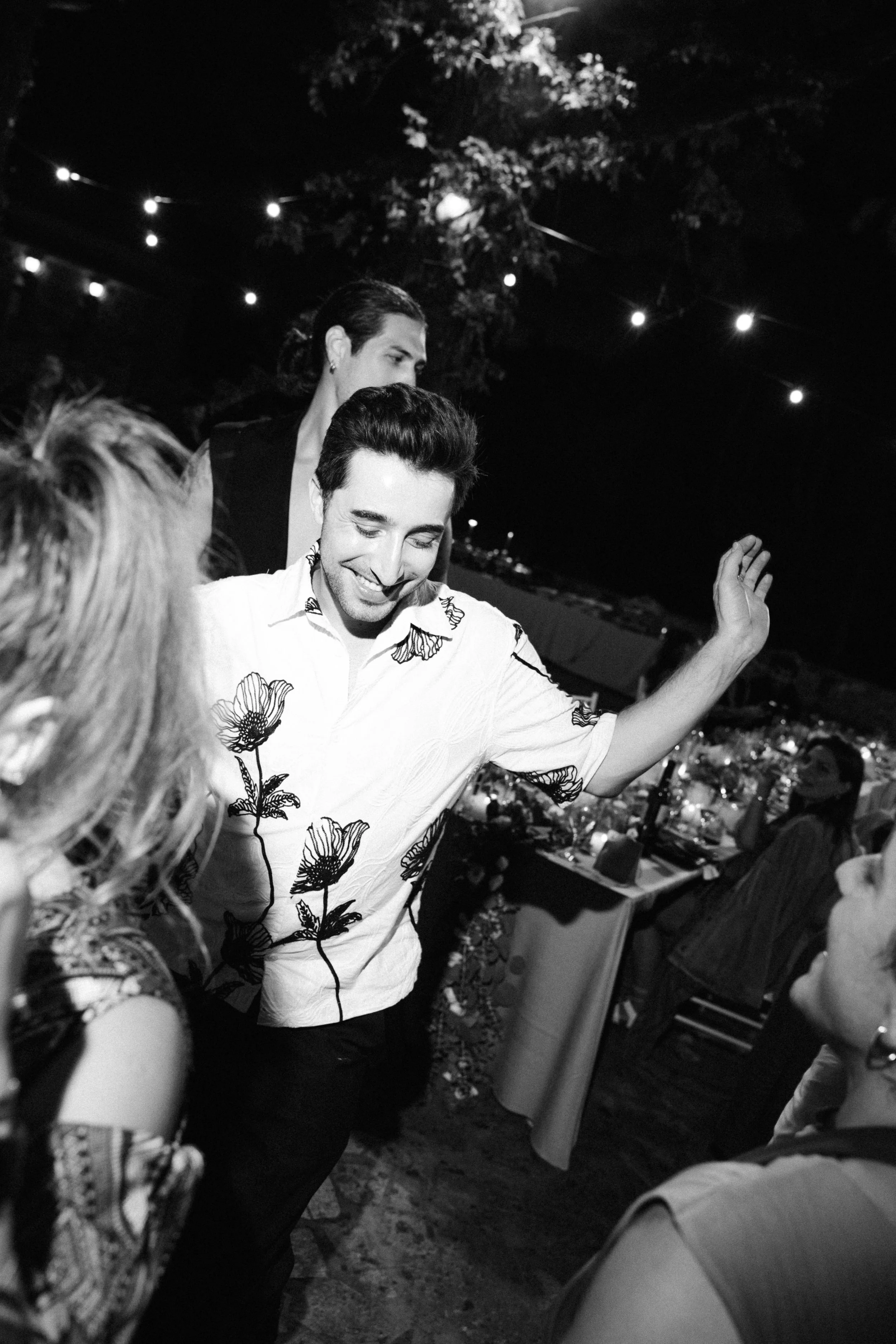 A man dancing at a nighttime outdoor party with string lights overhead, surrounded by women, some seated at a table with food and drinks.