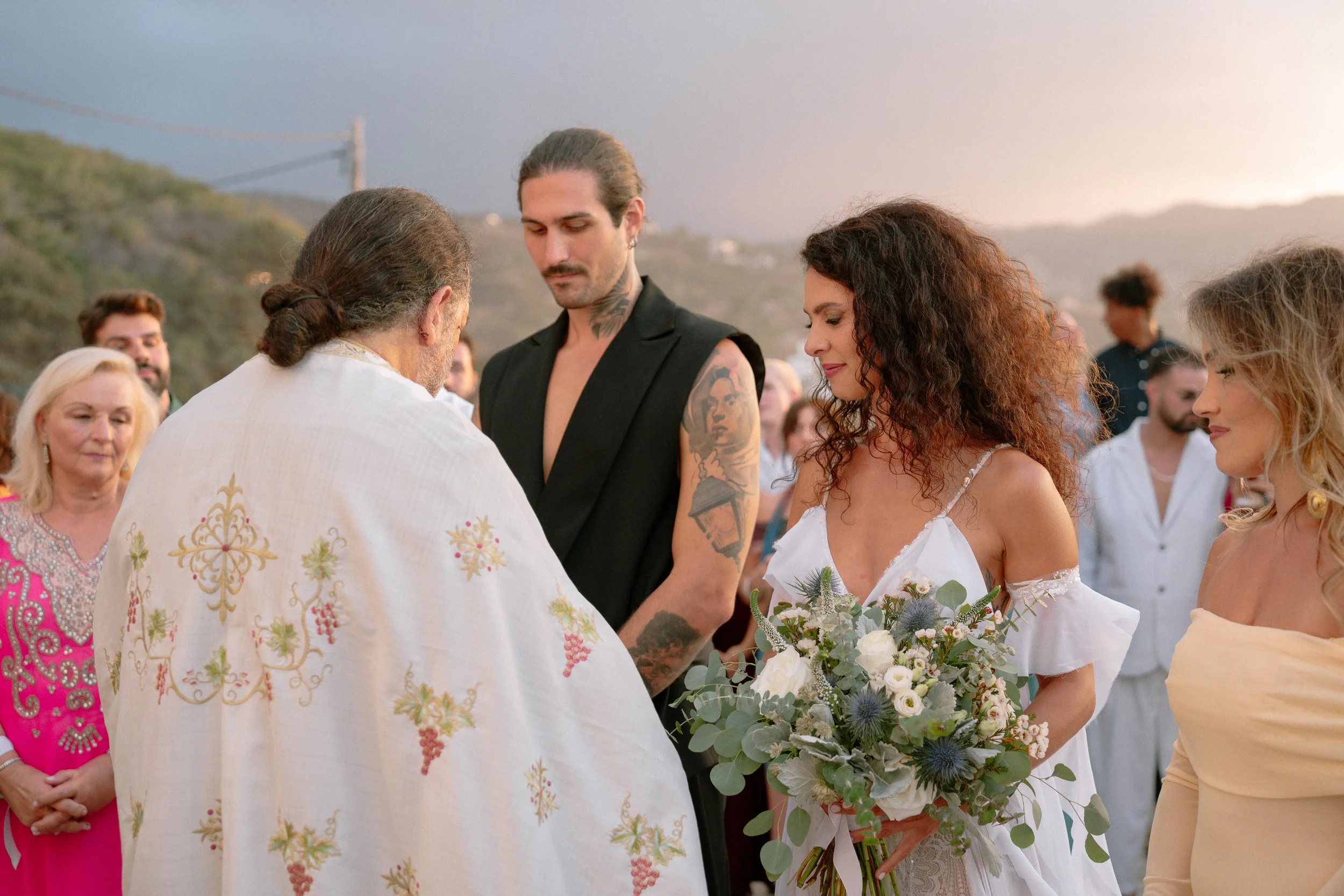 A wedding ceremony outdoors with a priest, a bride holding a bouquet, and a groom with tattoos, surrounded by guests.