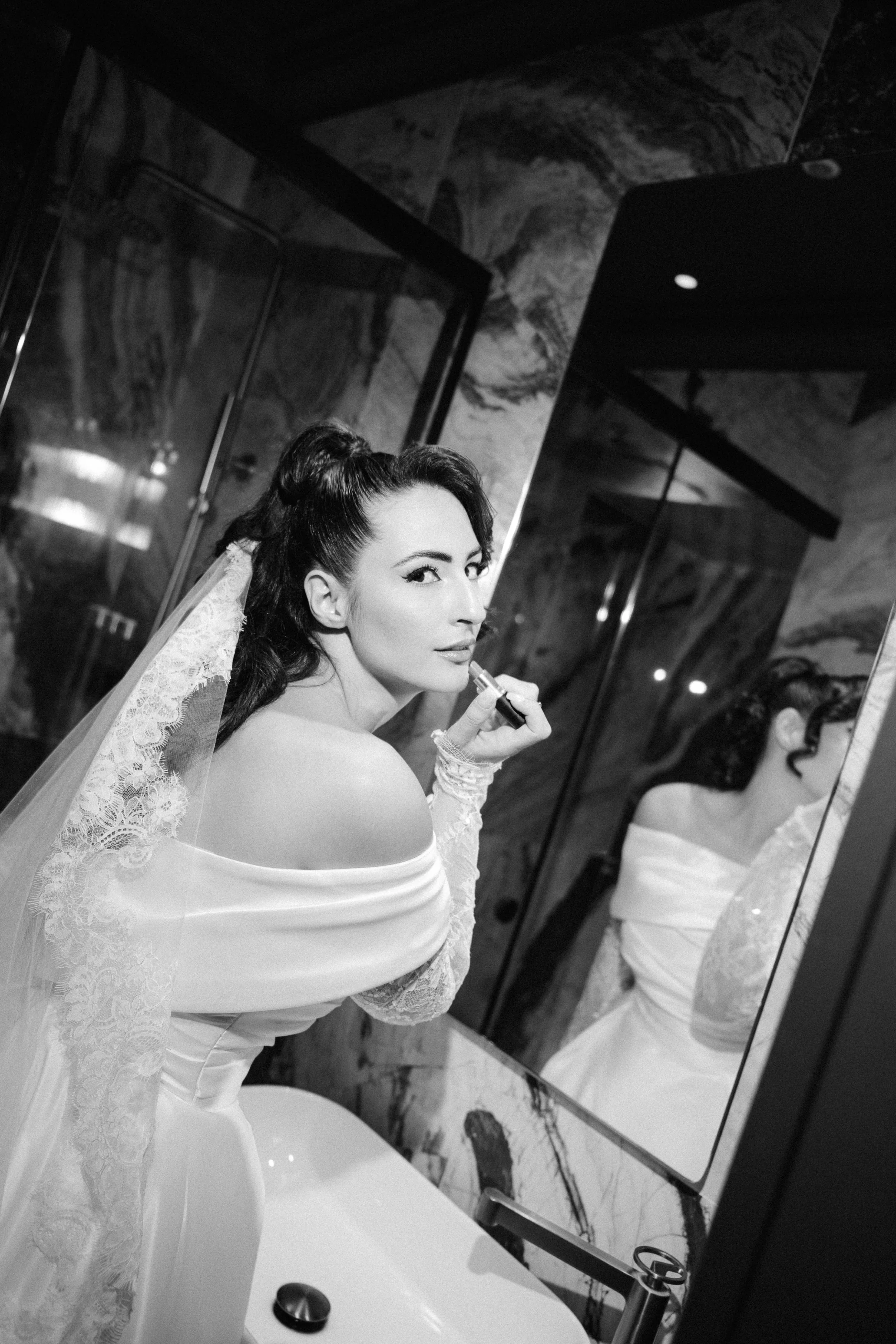 A bride in a wedding dress and lace veil looks into a mirror while applying lipstick in a hotel bathroom.