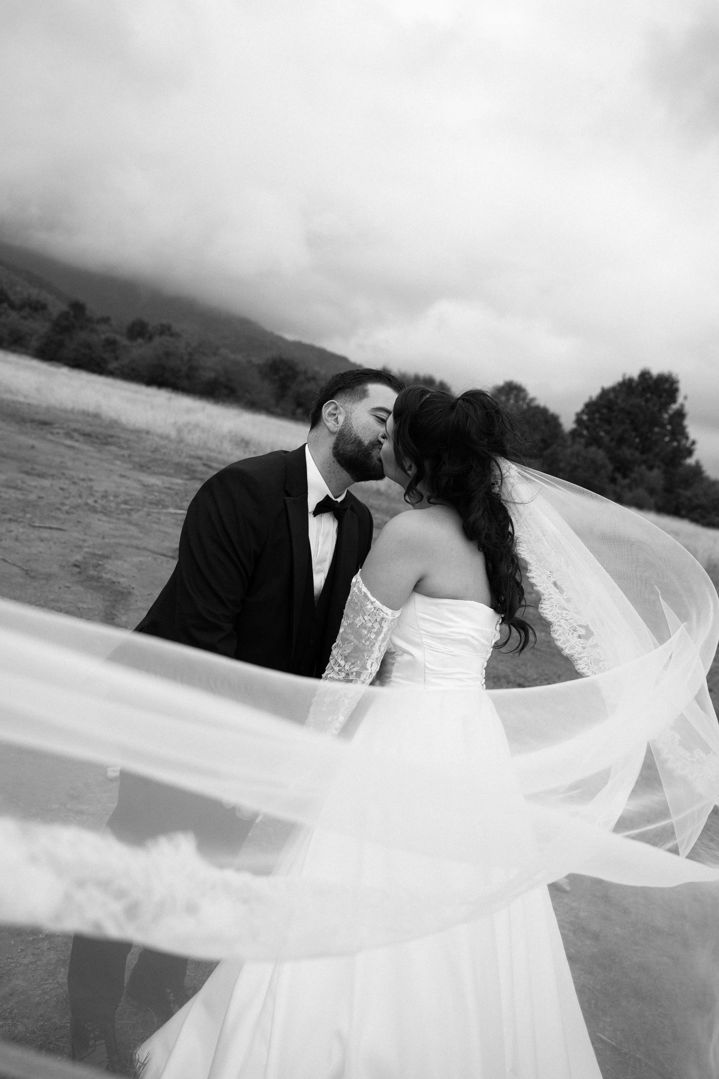 Black and white photo of a bride and groom kissing outdoors, mountains and trees in the background.