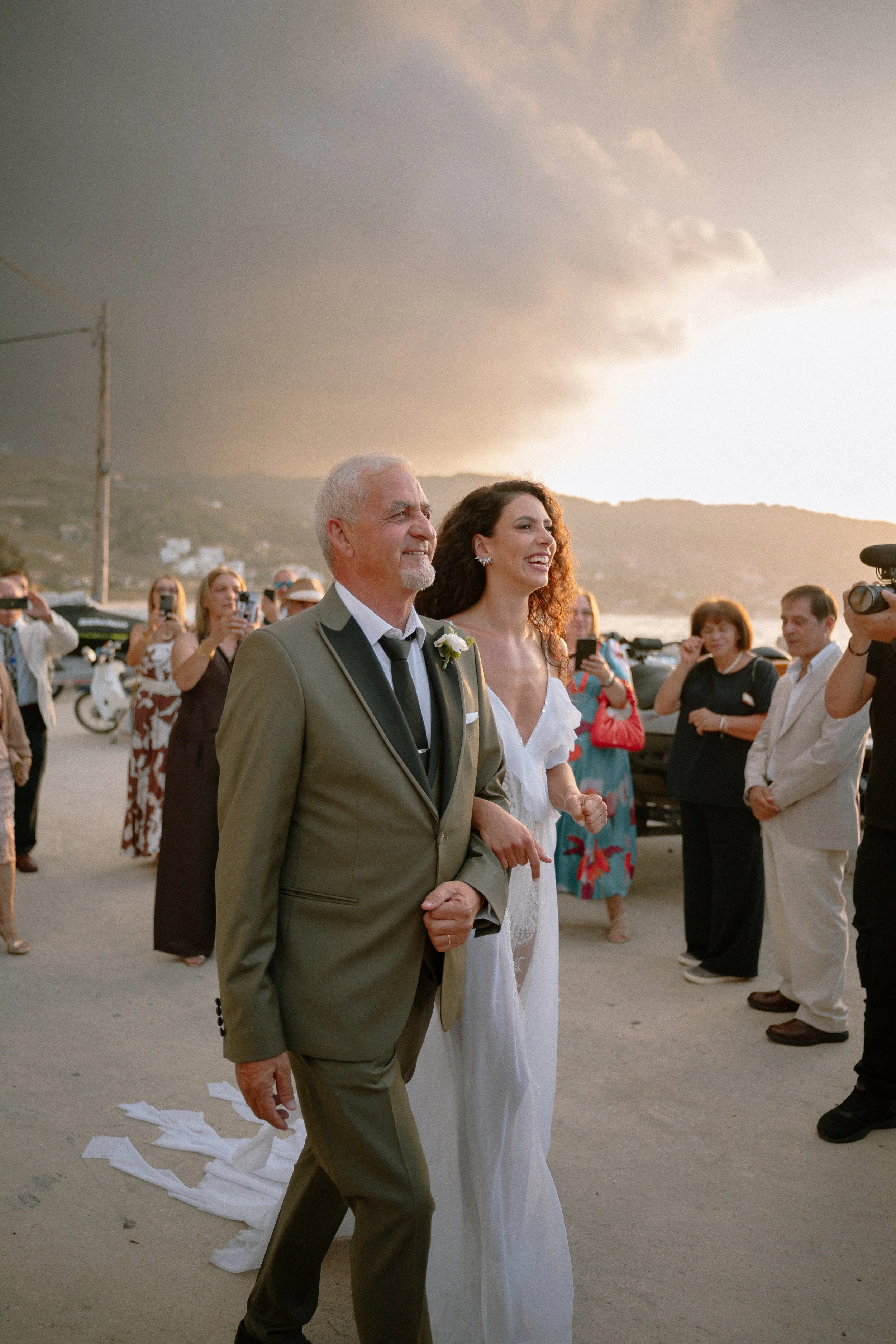 A bride walking down the aisle with an older man, likely her father, during a wedding ceremony outdoors at sunset, with guests taking photos around them and dark clouds in the sky.
