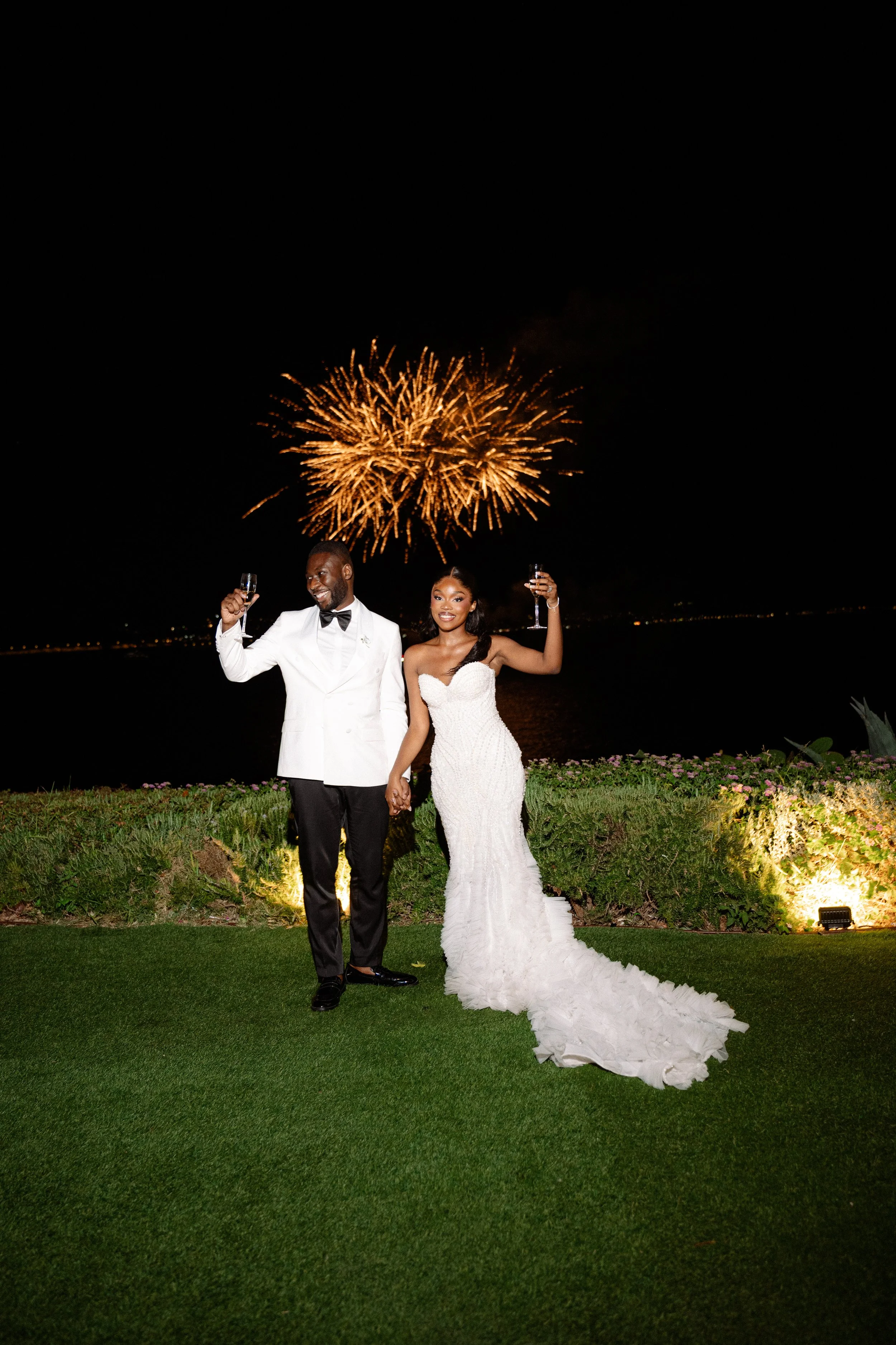A newlywed couple celebrating at night with fireworks in the sky, standing on grass near water, holding champagne glasses and smiling.