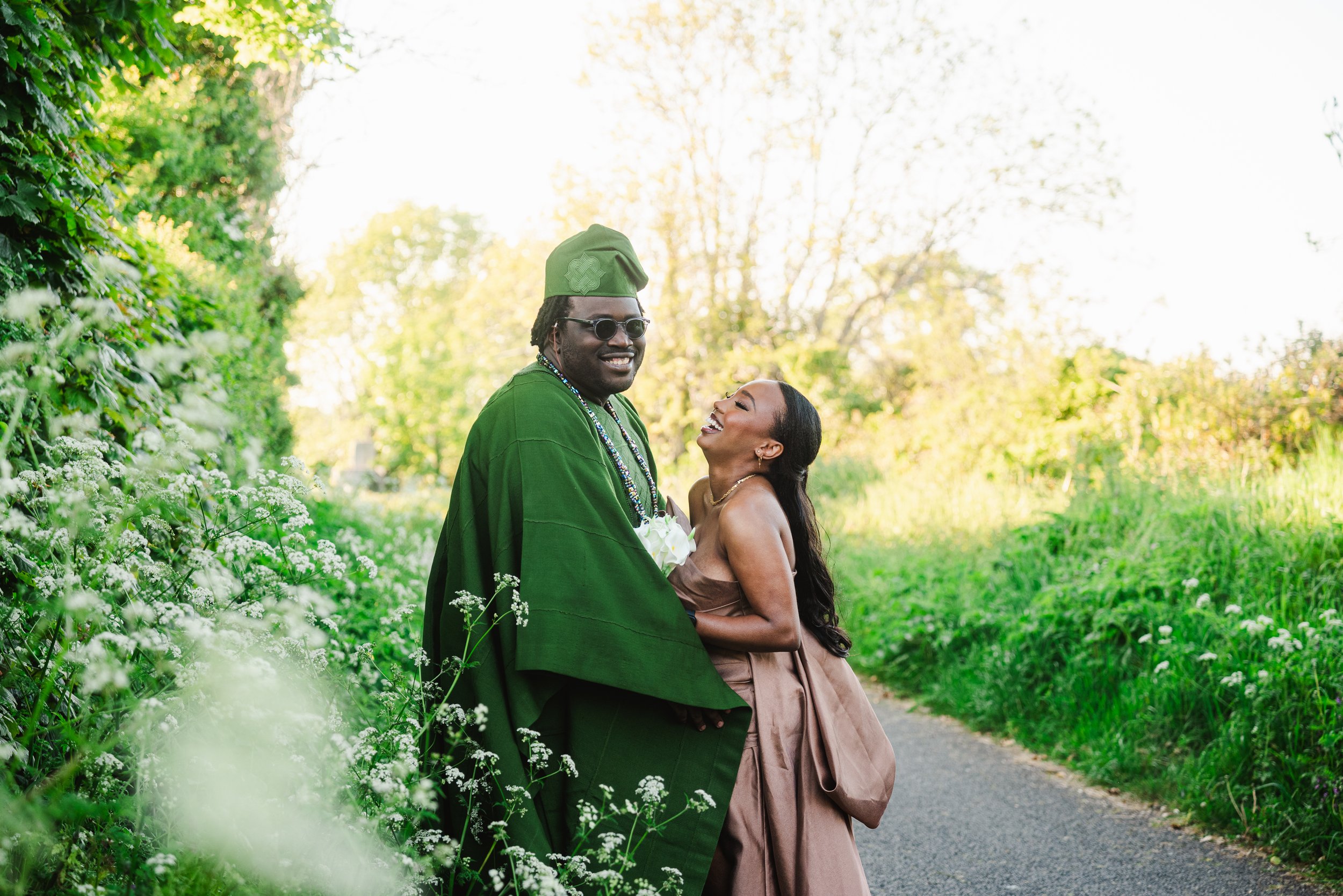 A couple dressed in formal attire stands outdoors on a path surrounded by greenery and white flowers, smiling and enjoying each other's company during a sunny day.