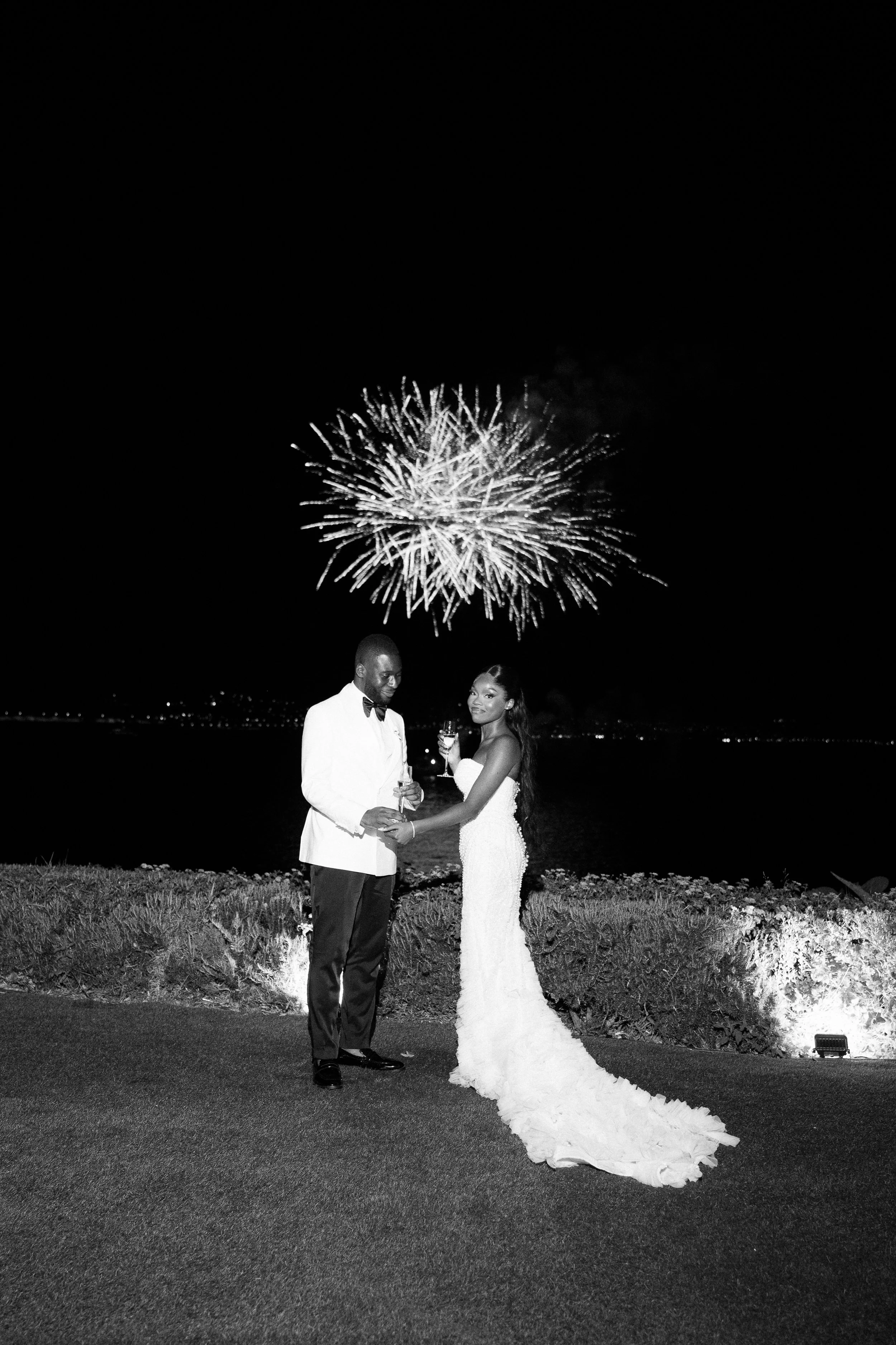 A bride and groom holding hands at night with fireworks in the sky behind them, standing near water and illuminated by landscape lighting.