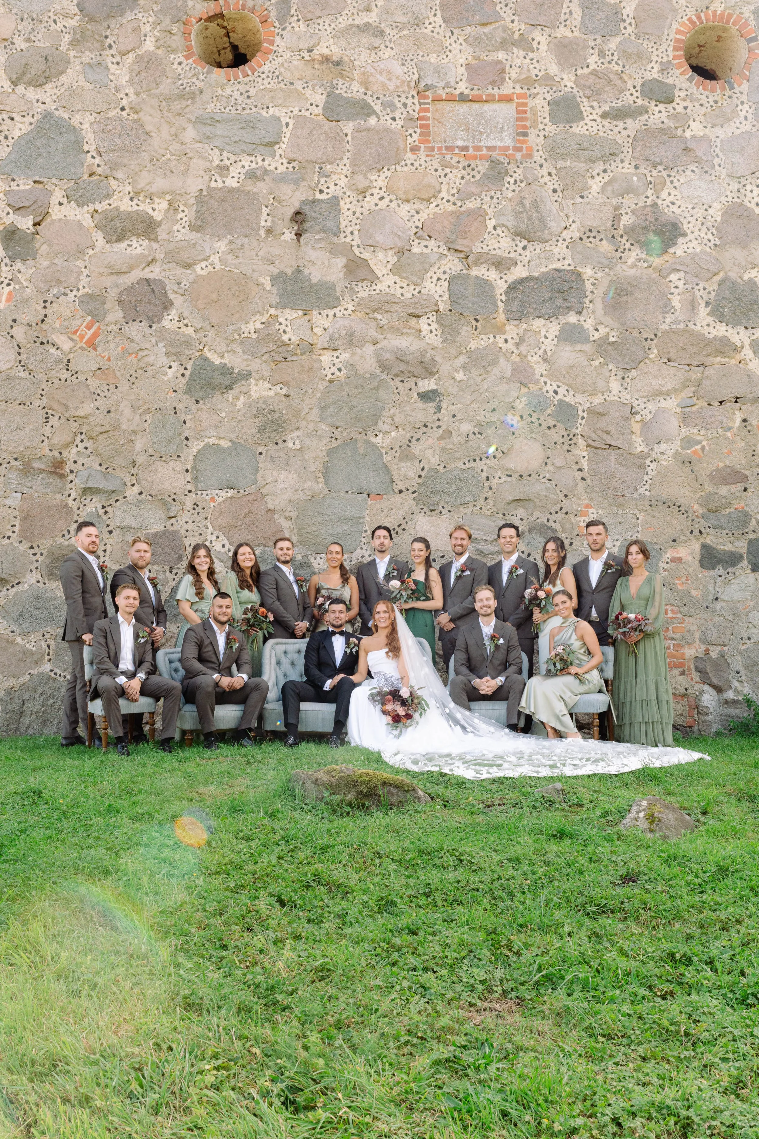 A wedding party with bride and groom sitting on a vintage sofa, surrounded by bridesmaids and groomsmen, in front of a large stone wall with small windows.