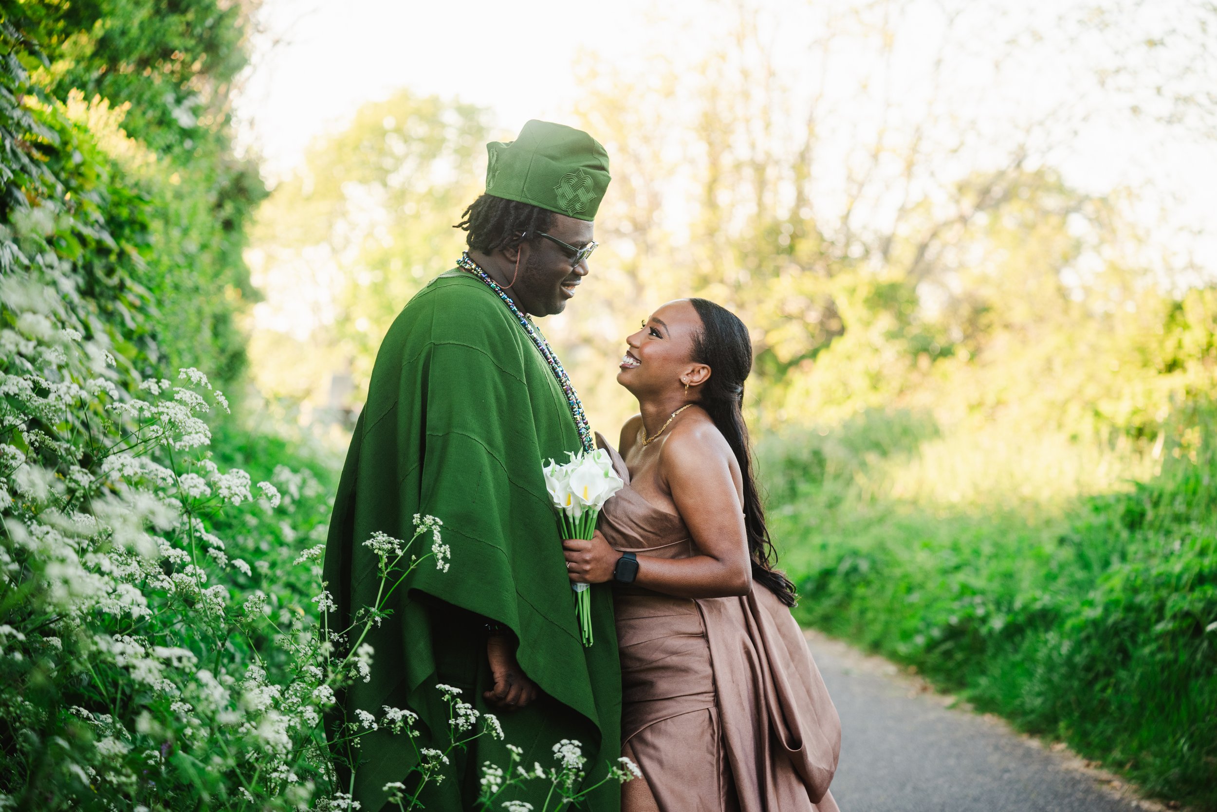 A couple stands close together outdoors on a path surrounded by greenery, with the man wearing green attire and the woman in a strapless brown dress holding a bouquet of white flowers, both smiling at each other.