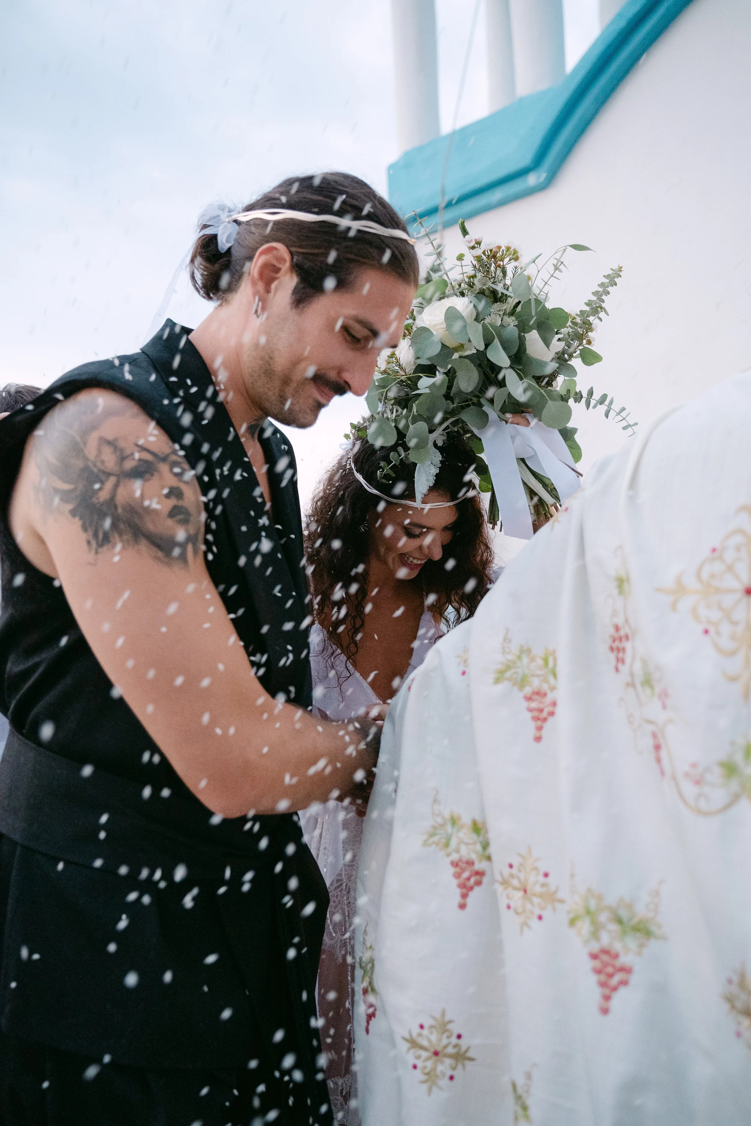 A couple getting married, standing close with a man with a tattooed arm, the woman has curly hair and a bouquet on her head, during a wedding ceremony with confetti falling around them.