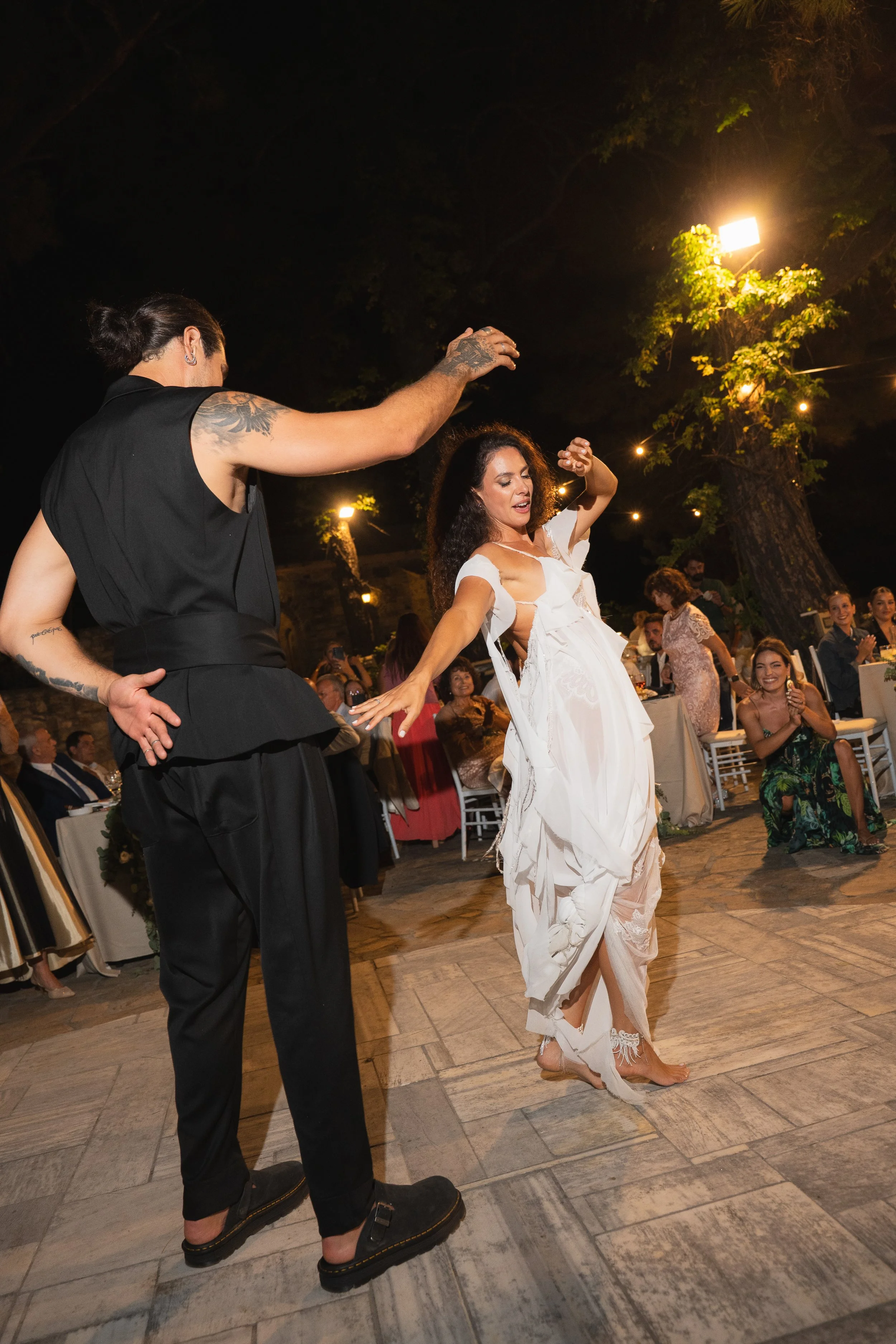 A woman dancing barefoot in a white dress at an outdoor night event, with a man in black clothing dancing nearby and an audience clapping and smiling around them.