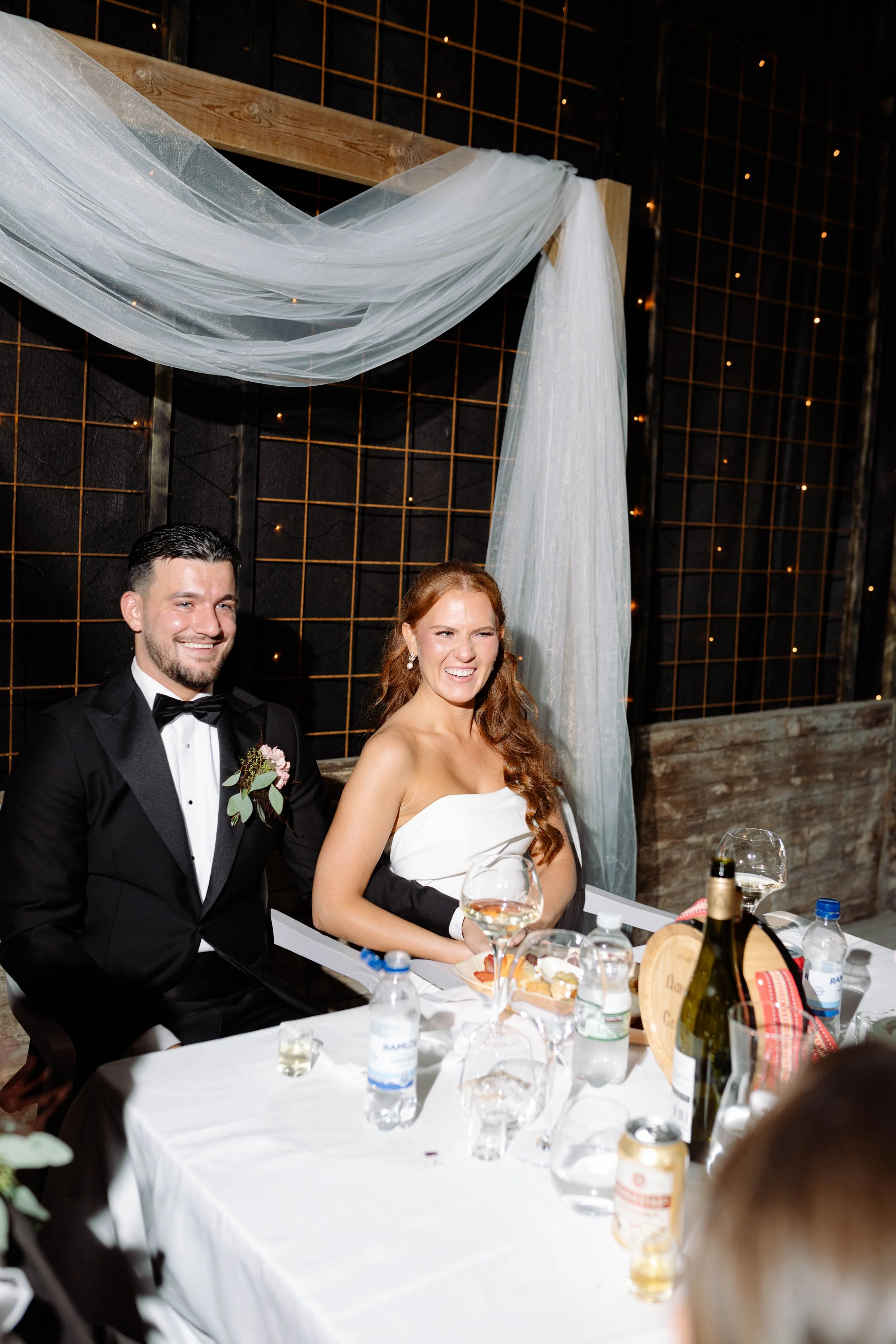 Bride and groom sitting at a table during a wedding reception, smiling, with drinks and food on the table.