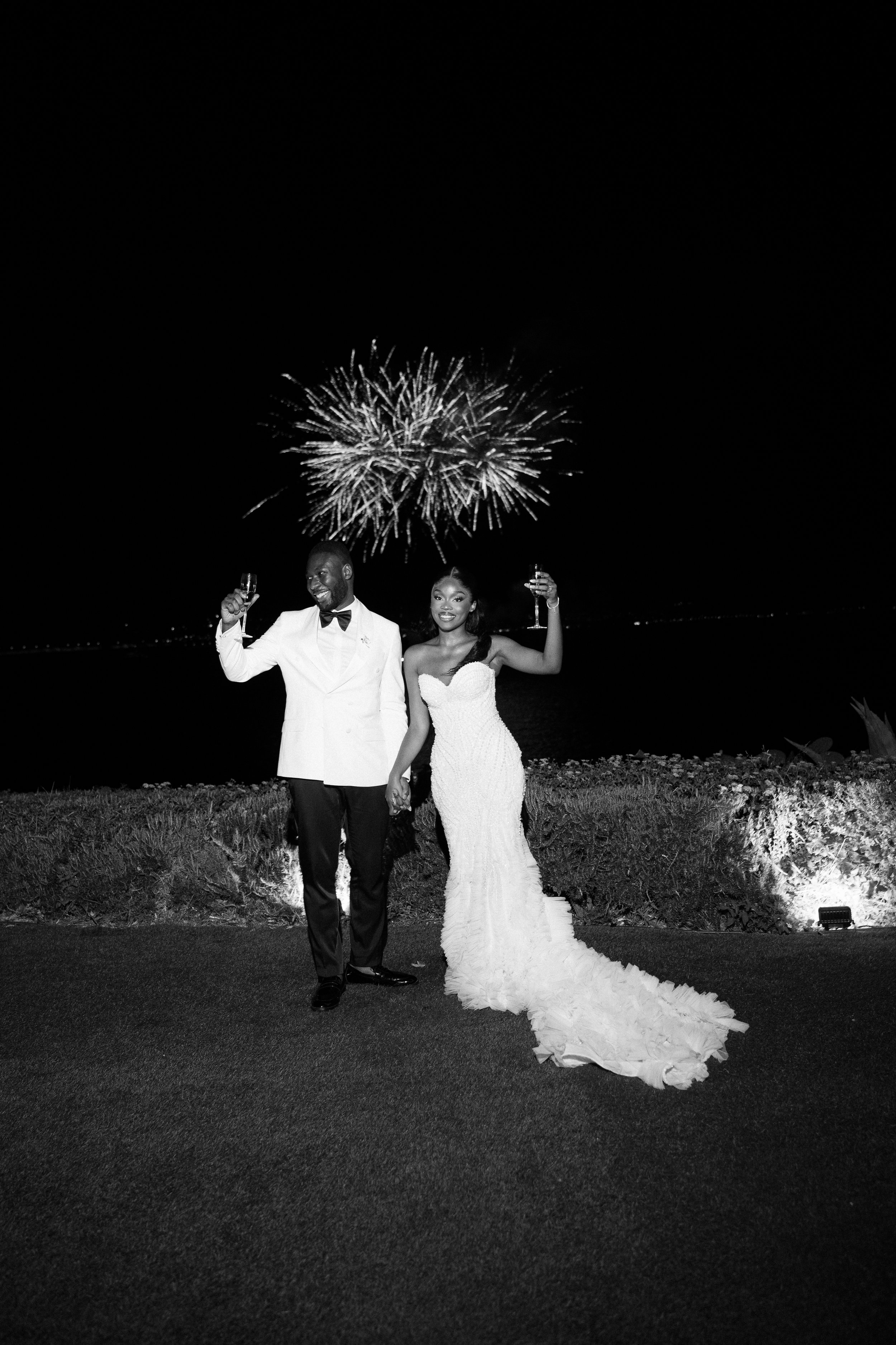 A black and white photo of a bride and groom celebrating with champagne during fireworks at night.