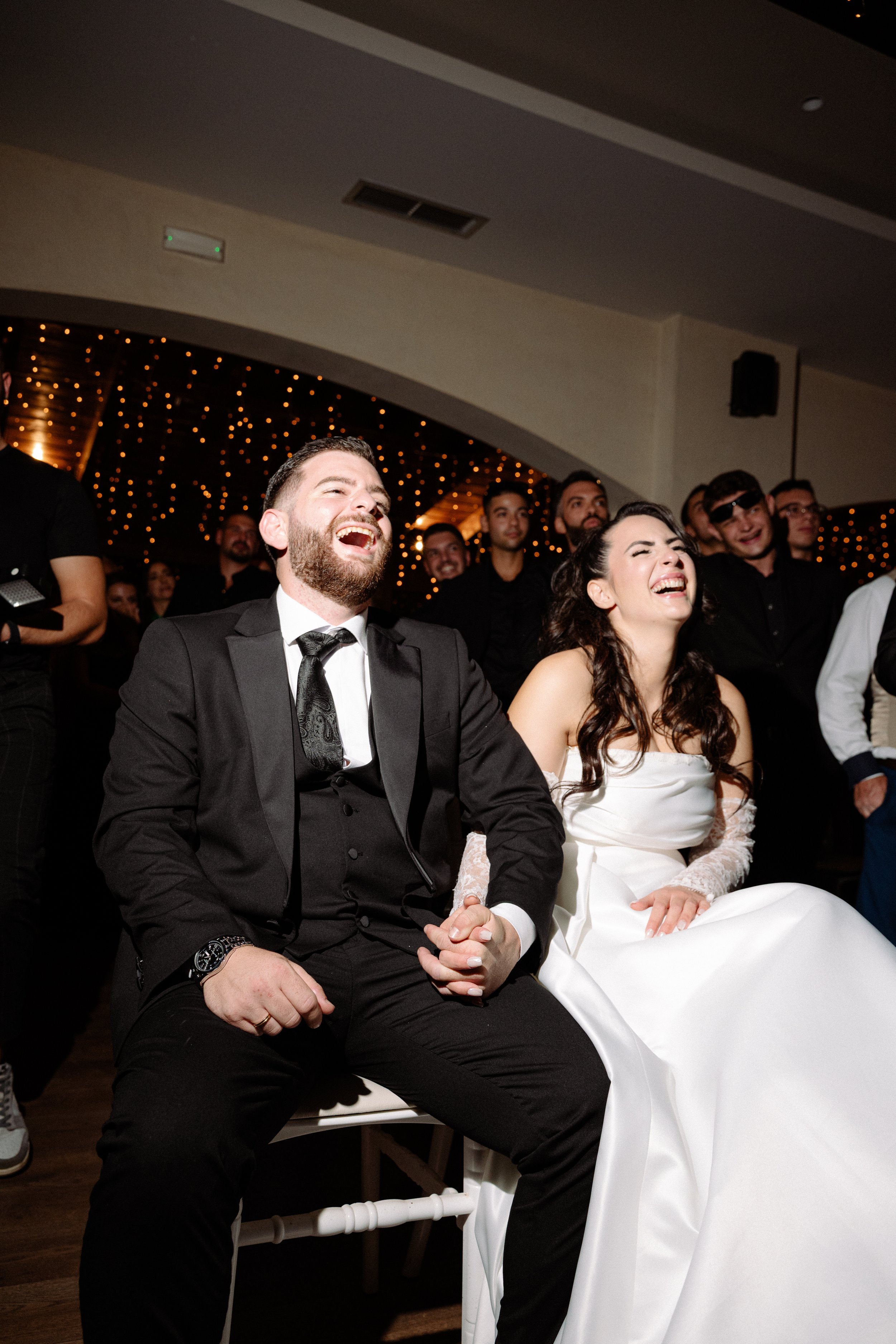 A wedding reception with a happy couple sitting together, laughing among guests, under warm string lights.