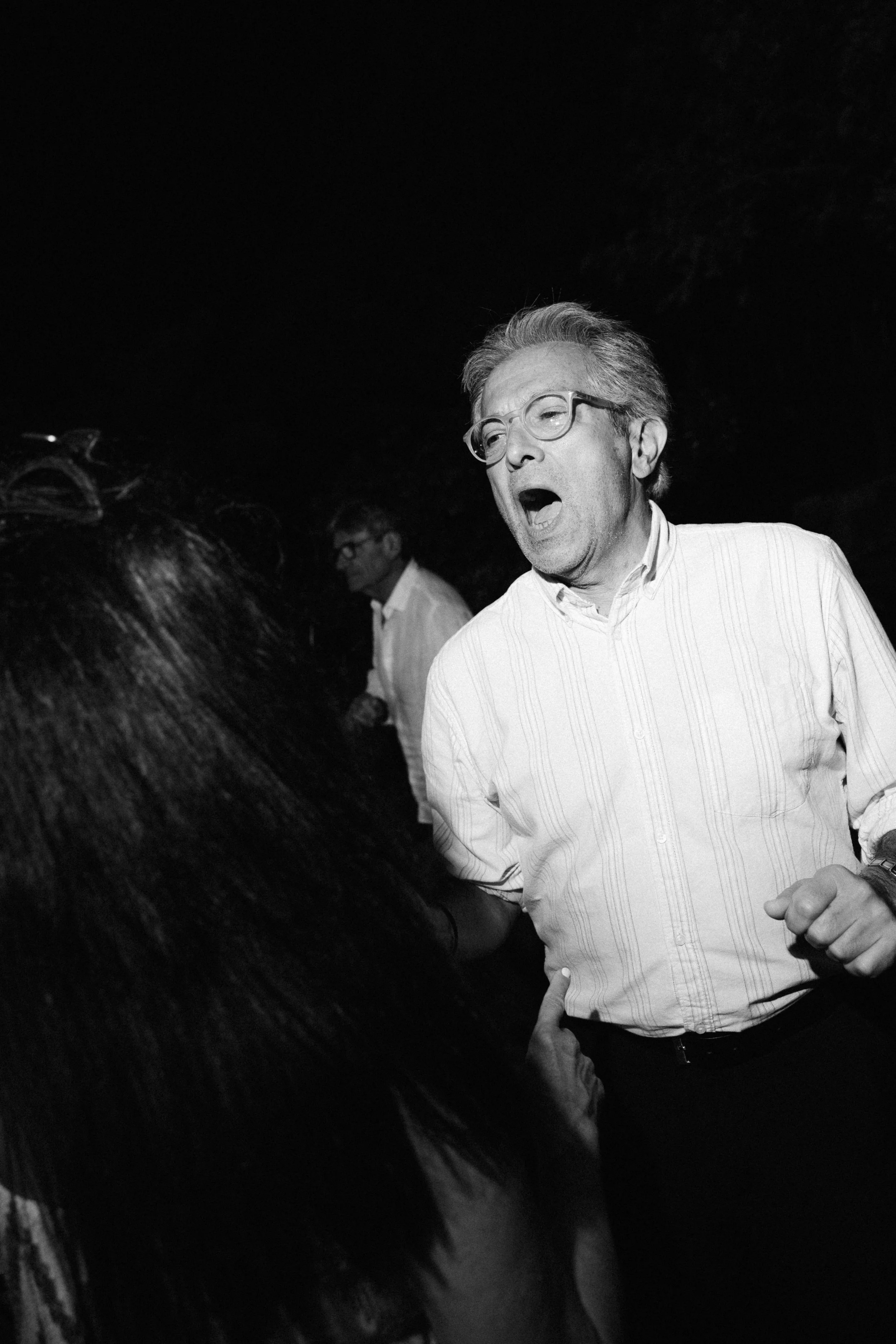 A man in glasses and a striped shirt appears to be shouting or singing, facing a woman with long dark hair, in a dark environment with another man in the background.