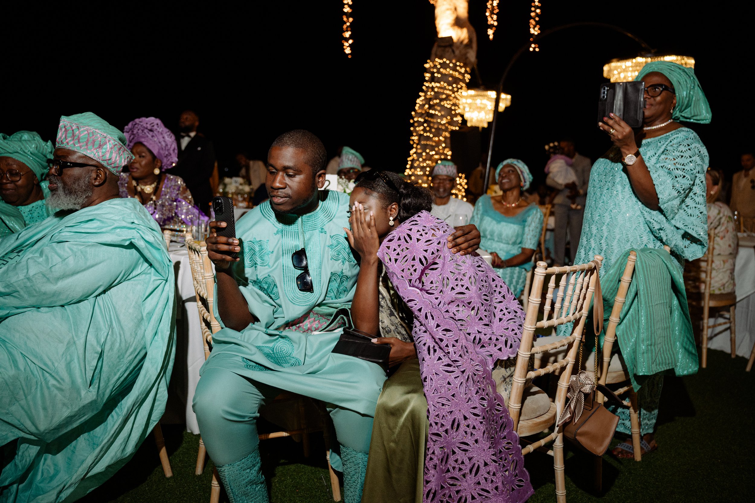 People dressed in traditional Nigerian clothing at an outdoor evening event with string lights in the background.