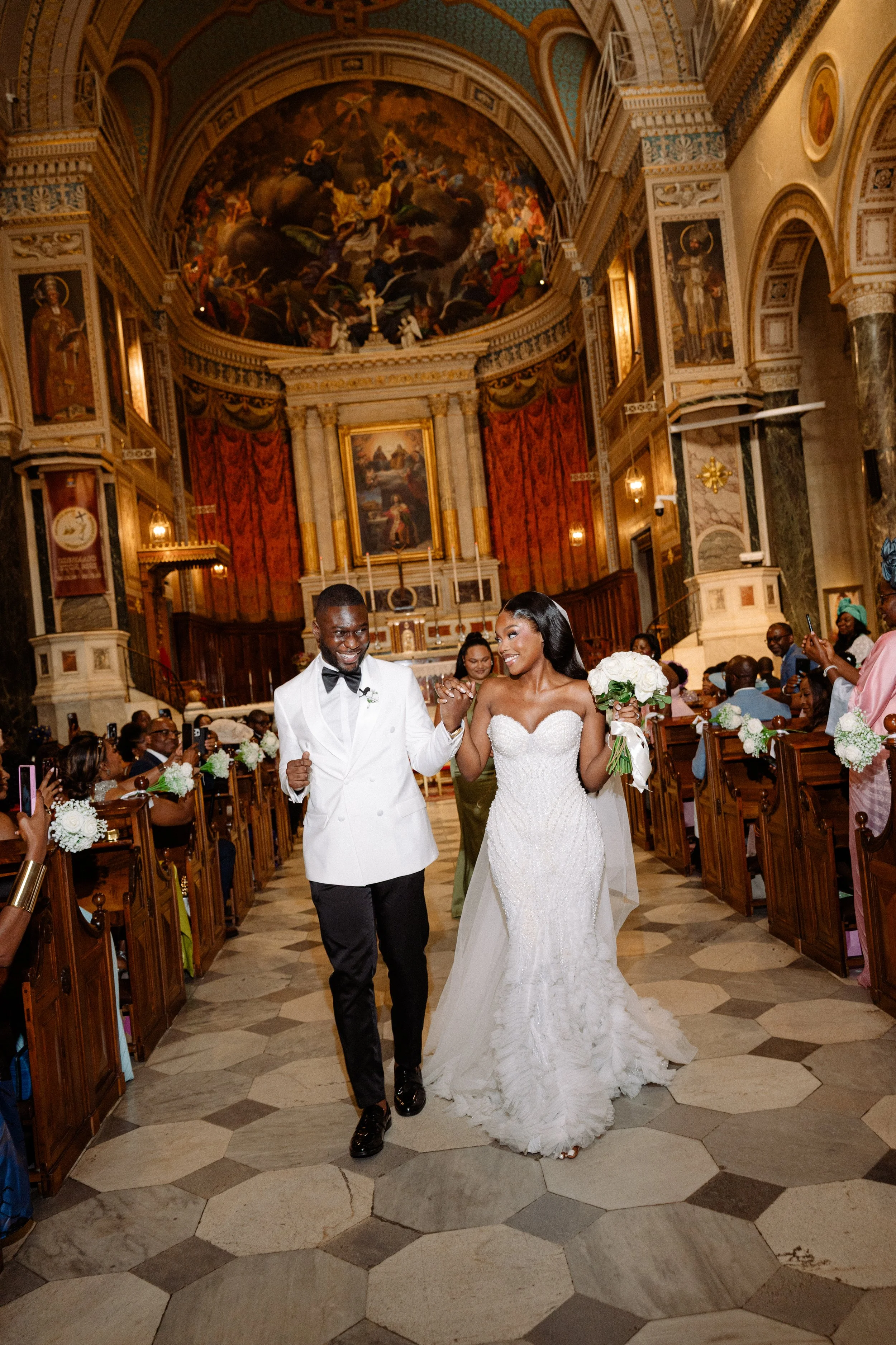 A bride and groom walking down the aisle in a church, smiling and holding hands, with guests in the background taking photos.
