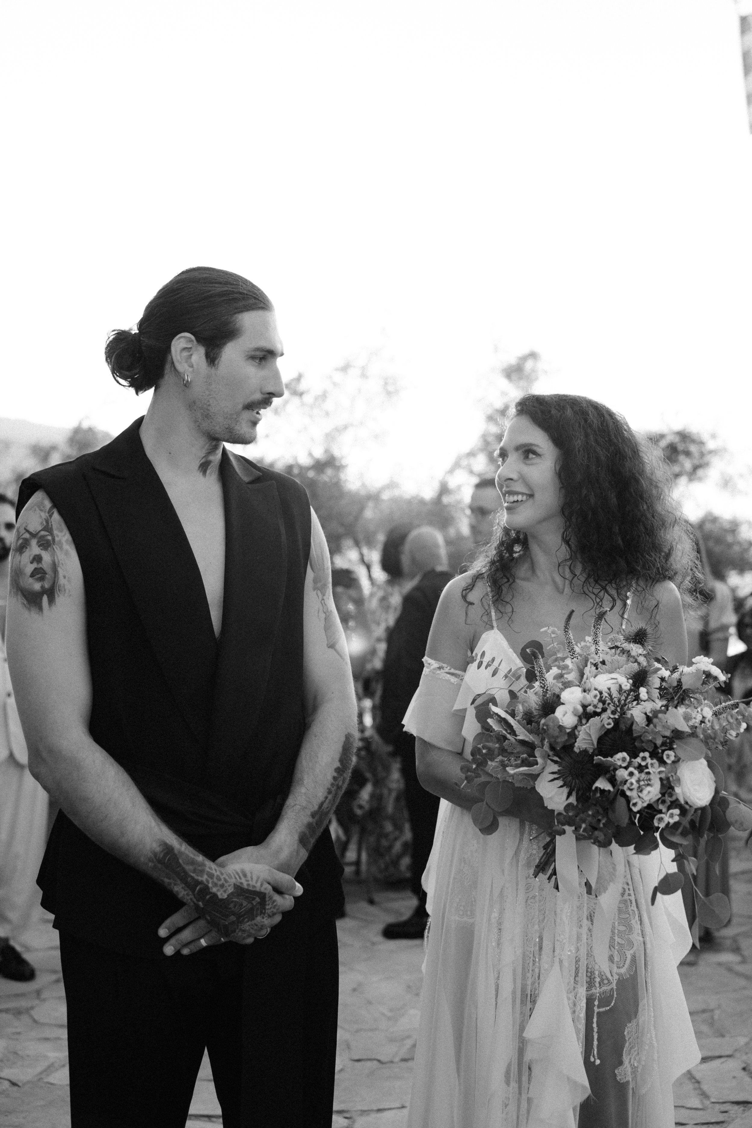 A black-and-white photo of a couple at a wedding, with the man having long hair pulled back and tattoos, and the woman holding a bouquet, exchanging glances.