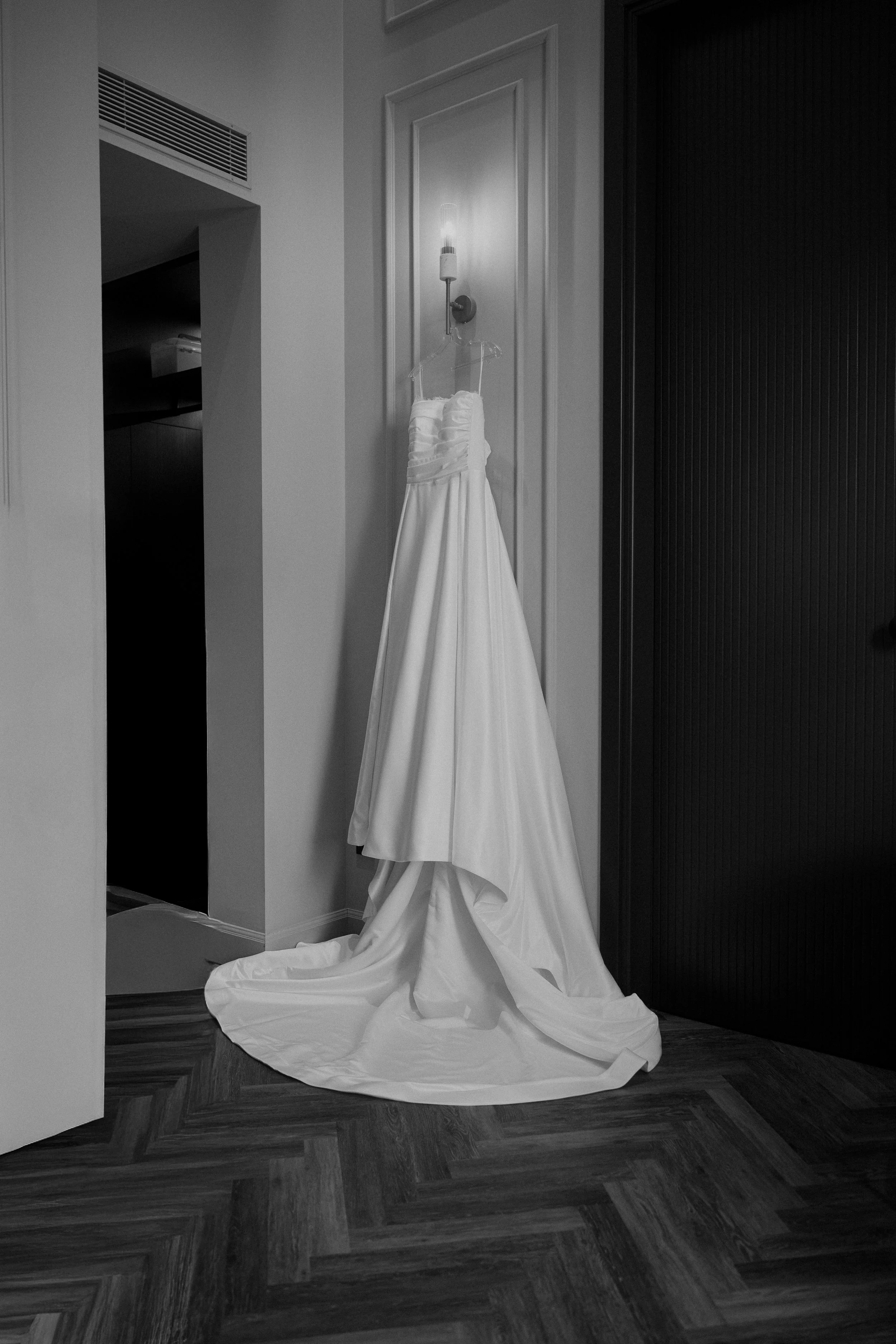 A wedding dress hanging on a hanger, draped over a wall hook with a wall sconce above it, on a wall next to a dark door panel.
